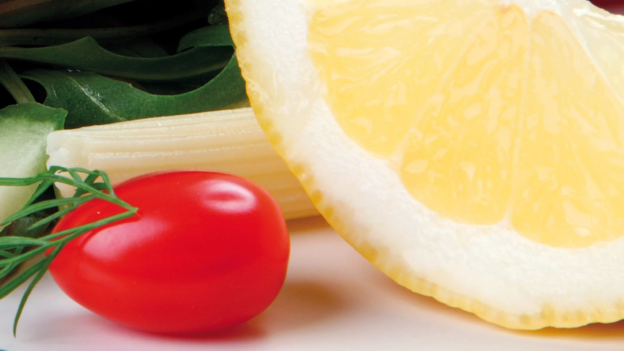 Close-up of fresh produce including a lemon slice, red cherry tomato, celery stalks, cucumber, and green leafy vegetables.