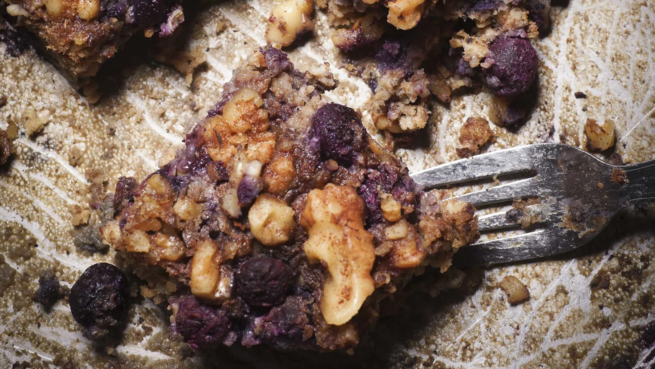 Close-up of a baked blueberry and walnut oatmeal bar on parchment paper with a fork resting beside it.