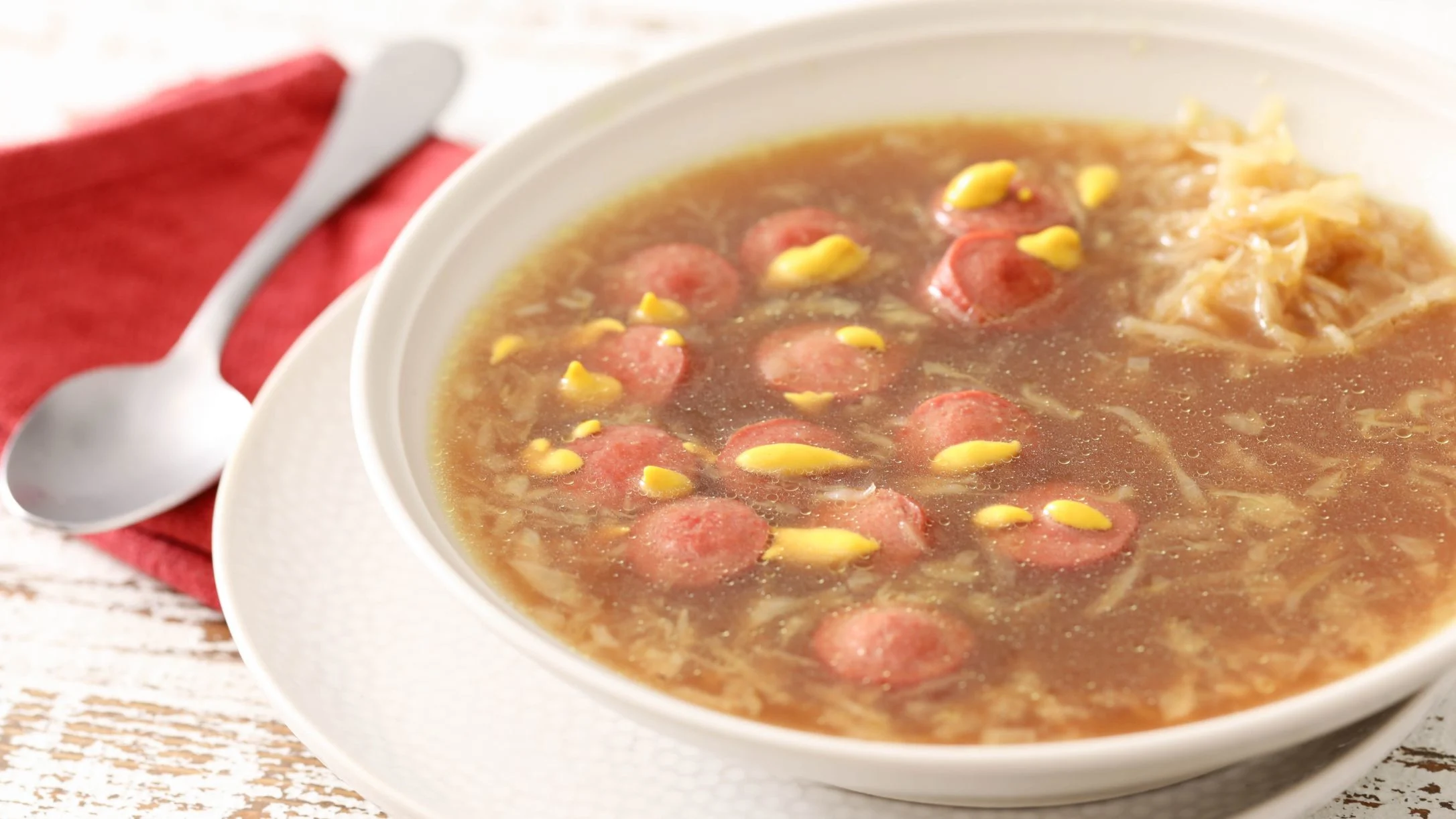 A white bowl of broth-based soup with sliced sausage, mustard drizzle, and sauerkraut, served with a spoon on a red napkin.