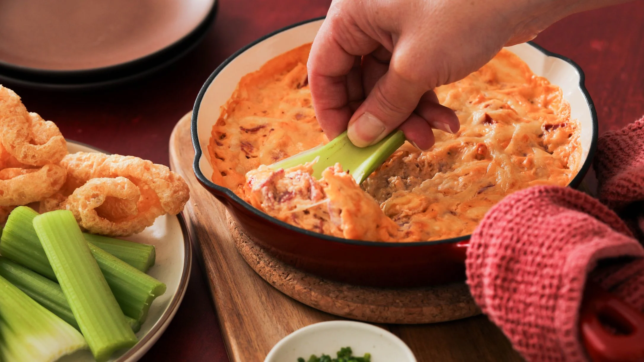 A hand dips a celery stick into a bubbling orange reuben dip in a cast iron dish, served with celery and pork rinds.