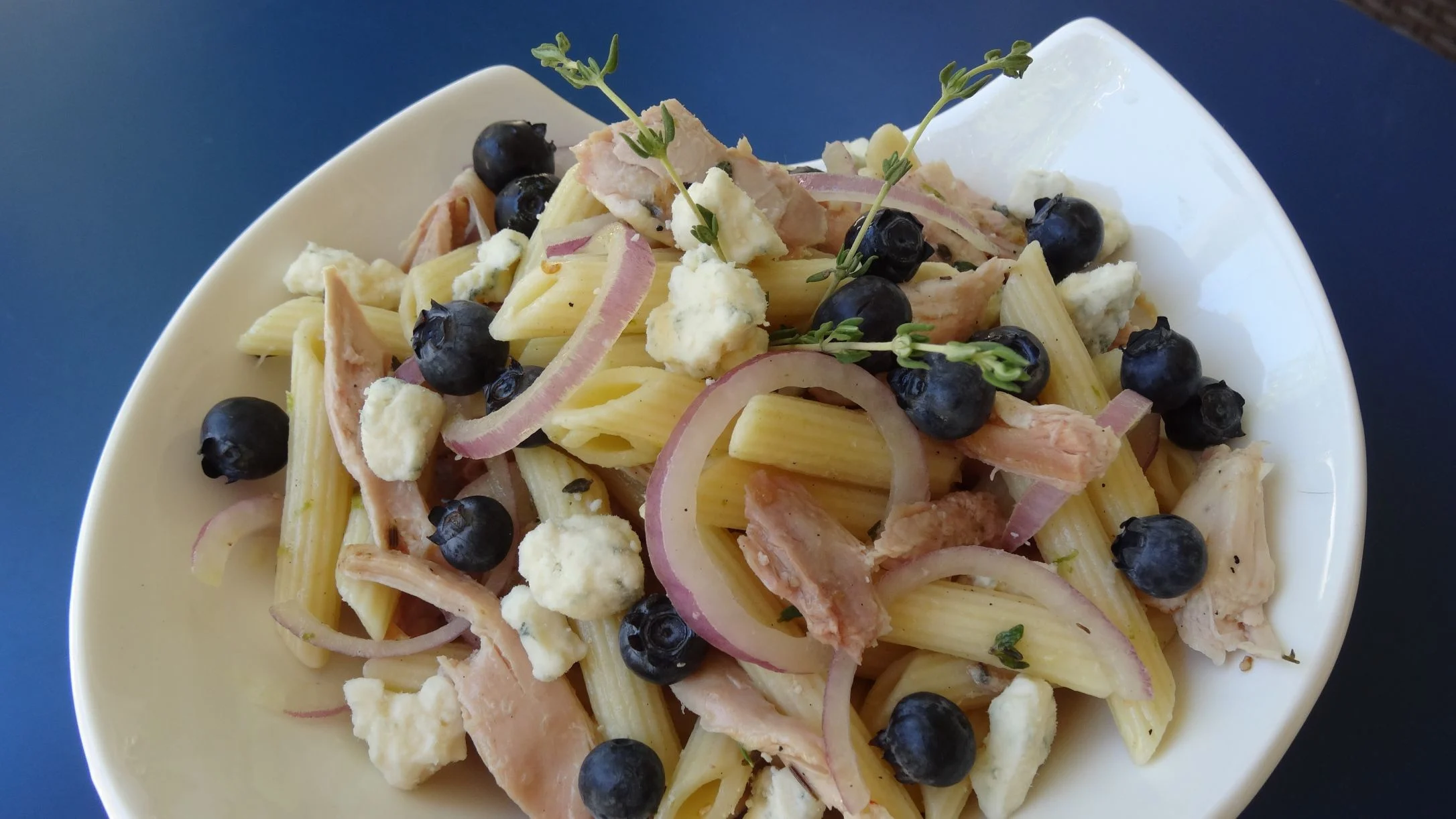 Penne pasta salad with blueberries, chicken, red onion, crumbled cheese, and fresh thyme on a white plate.