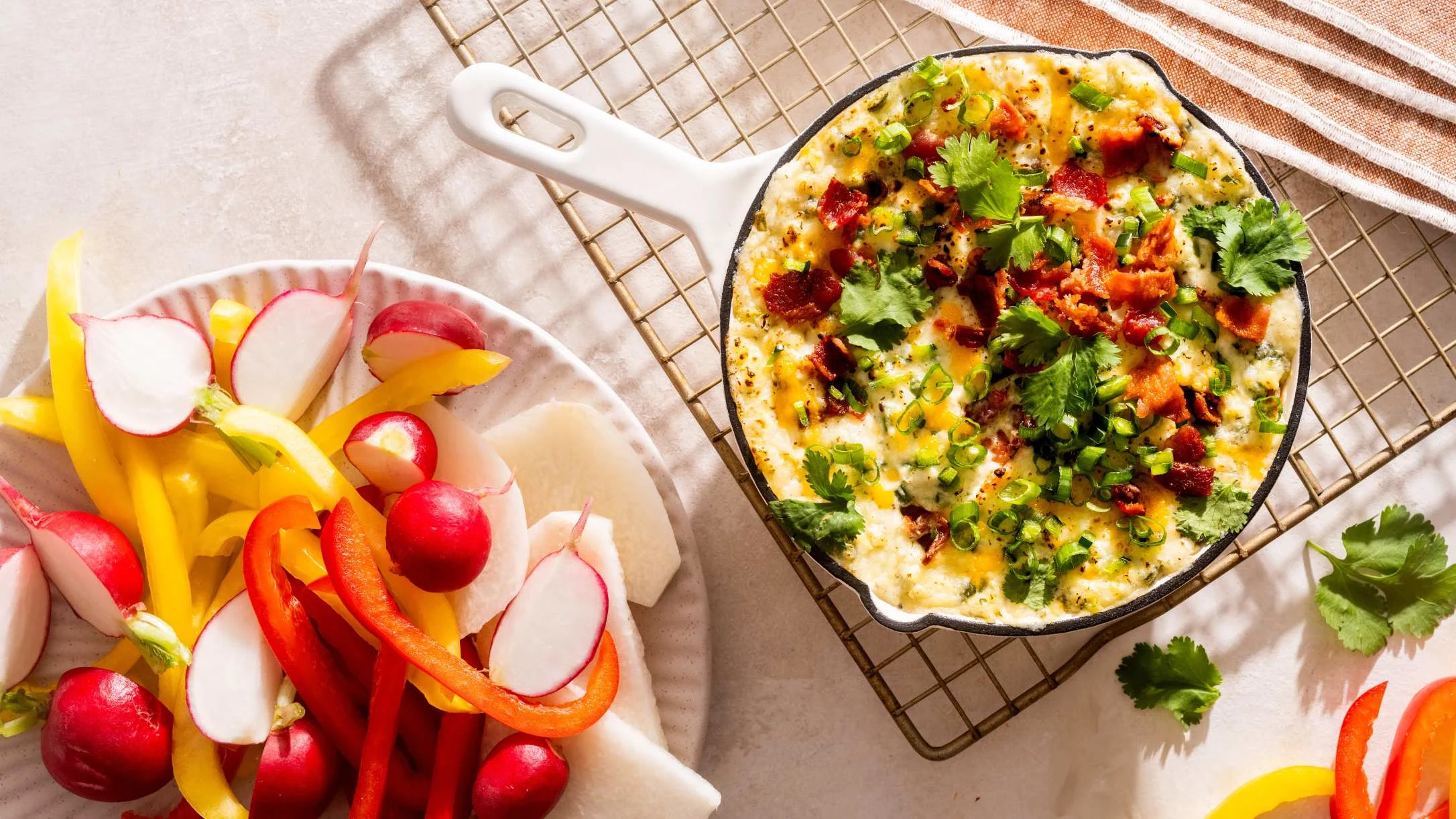 Cheesy dip topped with bacon, cilantro, and green onions in a cast iron skillet, served with sliced peppers and radishes.