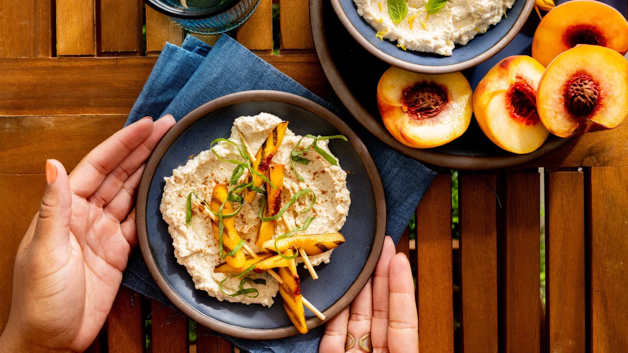 Two hands holding a plate of grilled peach skewers on creamy ricotta, with fresh peach halves nearby on a wooden table.