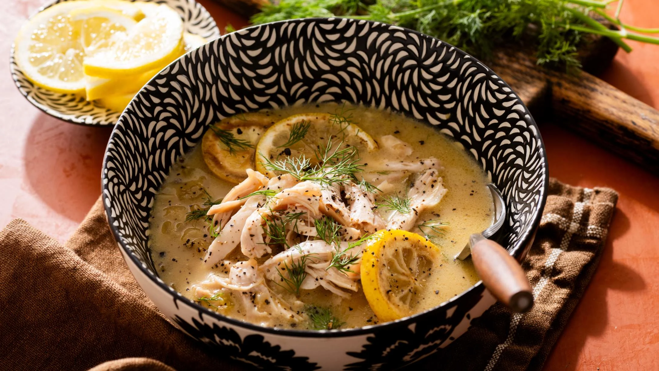 A bowl of creamy chicken soup with lemon slices, fresh dill, and black pepper in a black-and-white patterned bowl.