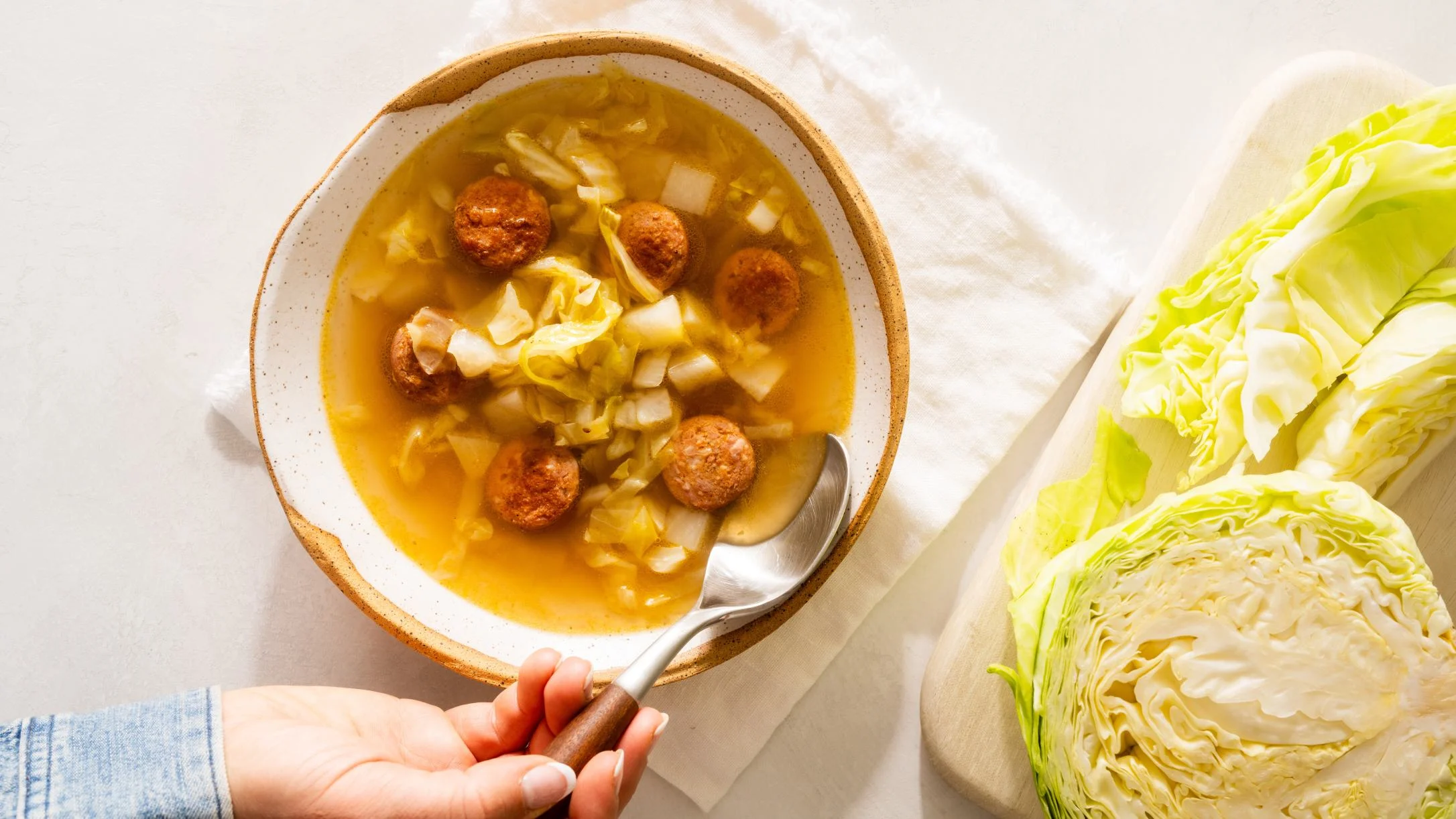 Bowl of cabbage soup with kielbasa being served, with fresh cabbage head on wooden board beside it.
