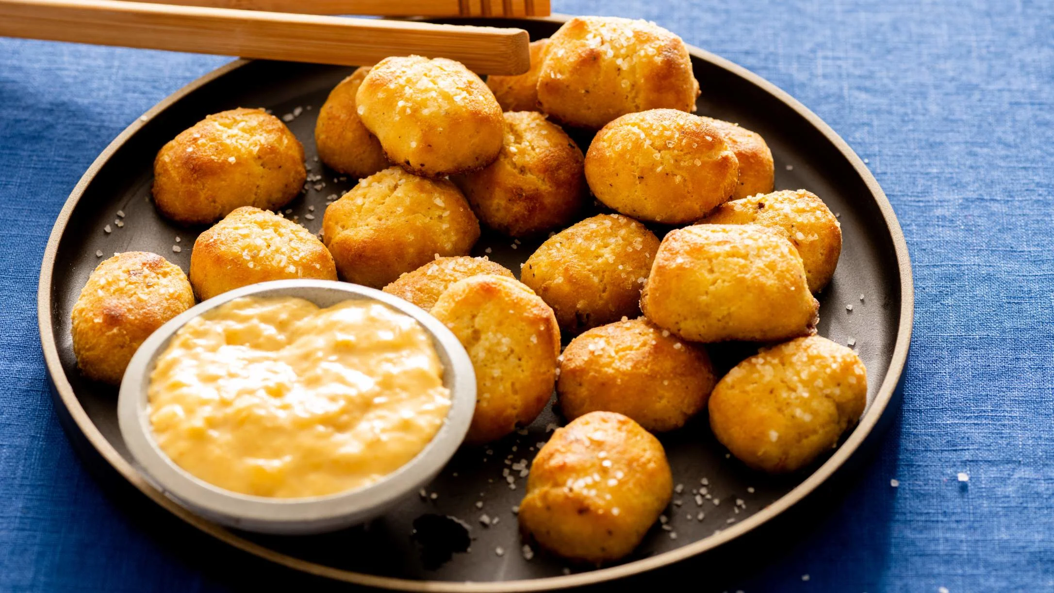 Golden pretzel bites sprinkled with salt on a dark plate, served with a creamy dipping sauce, on a blue linen surface.