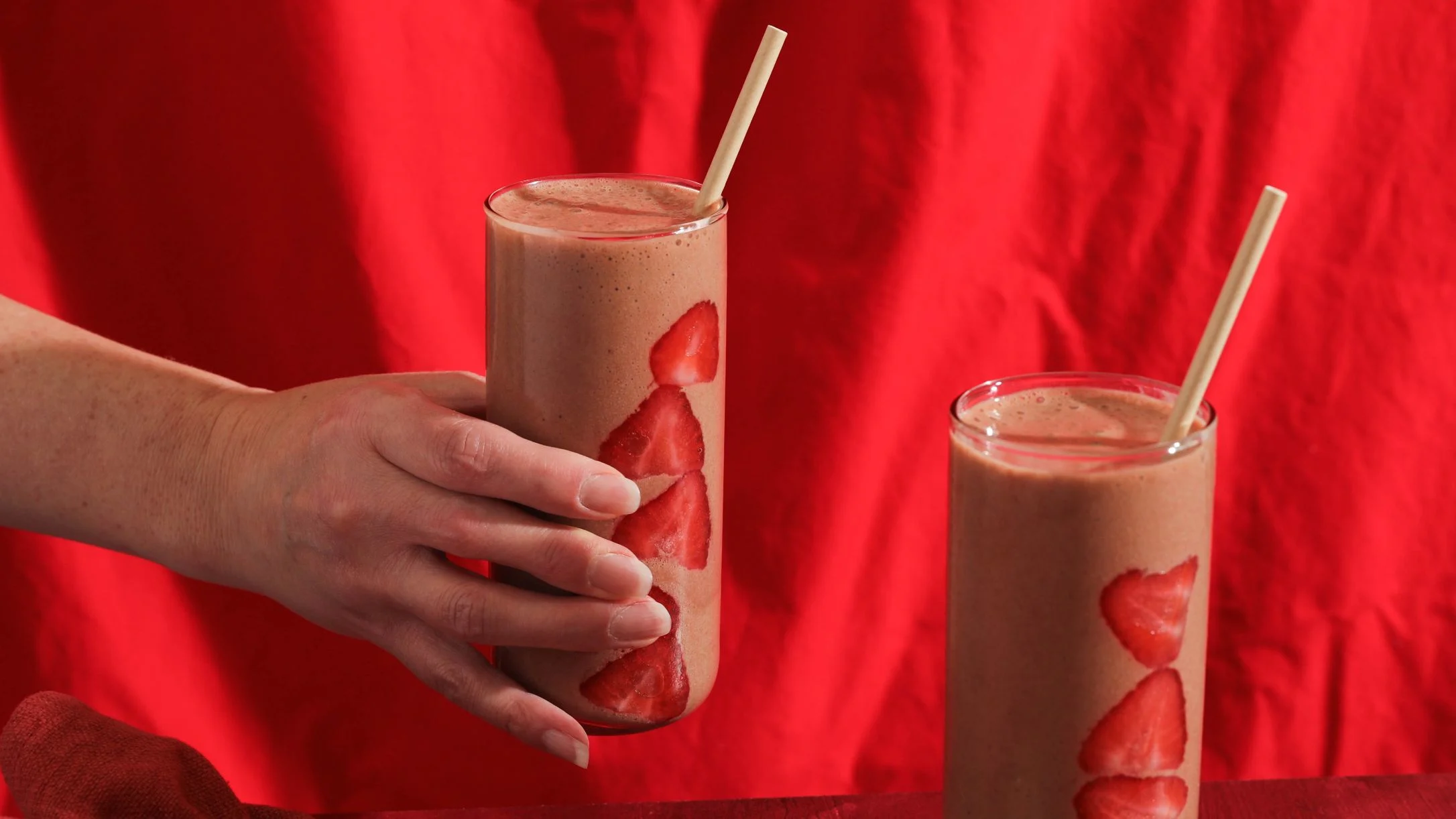 A hand holds a strawberry smoothie with sliced strawberries pressed against the glass and a straw, beside a second smoothie on a red background.