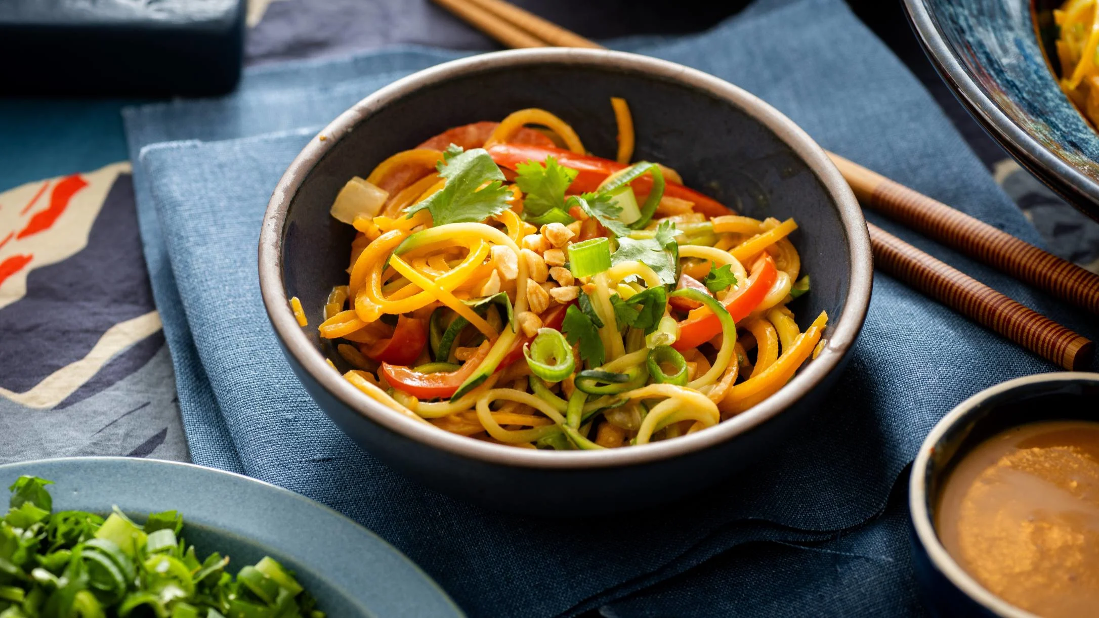 A dark bowl of colorful vegetable noodles topped with cilantro, peanuts, and green onions, with chopsticks and dipping sauce nearby.