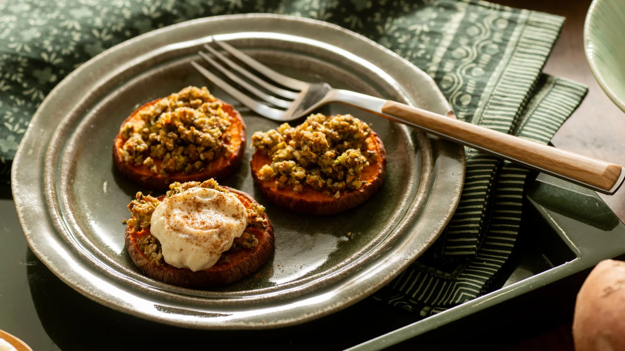 Three roasted sweet potato rounds topped with nut crumble and cream on a rustic metal plate with a fork.