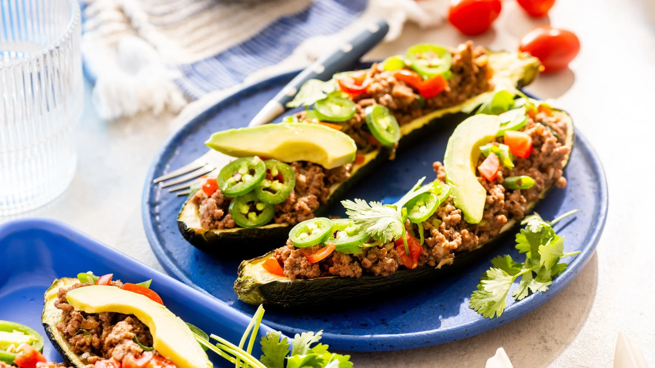 Stuffed zucchini halves filled with seasoned ground meat, topped with jalapeños, tomatoes, and avocado slices on a blue plate.