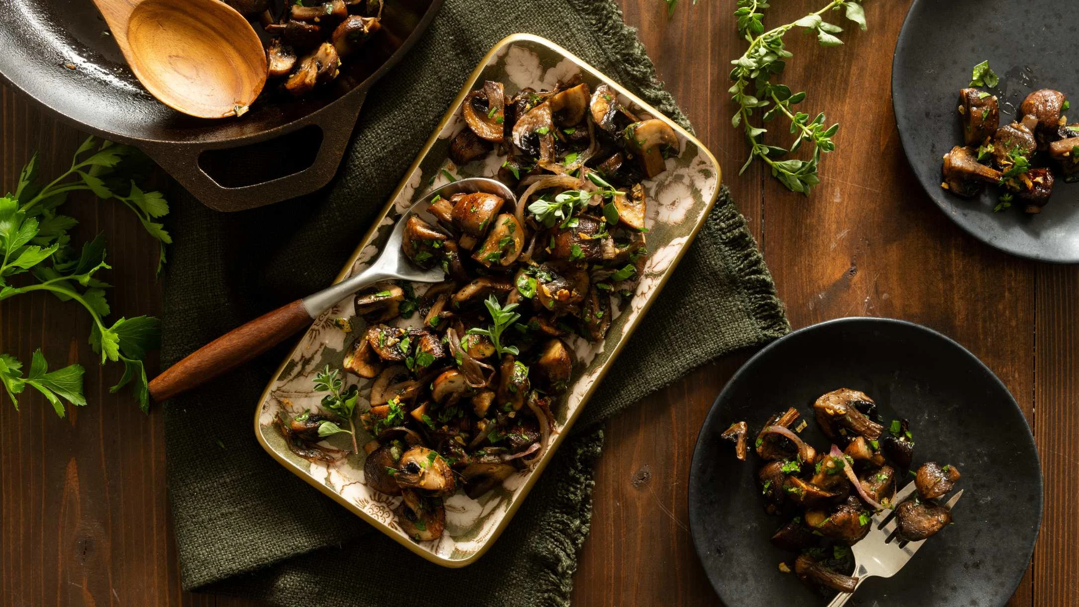 Sautéed mushrooms with fresh herbs on a decorative platter, cast iron skillet, and dark plates on a wooden table.