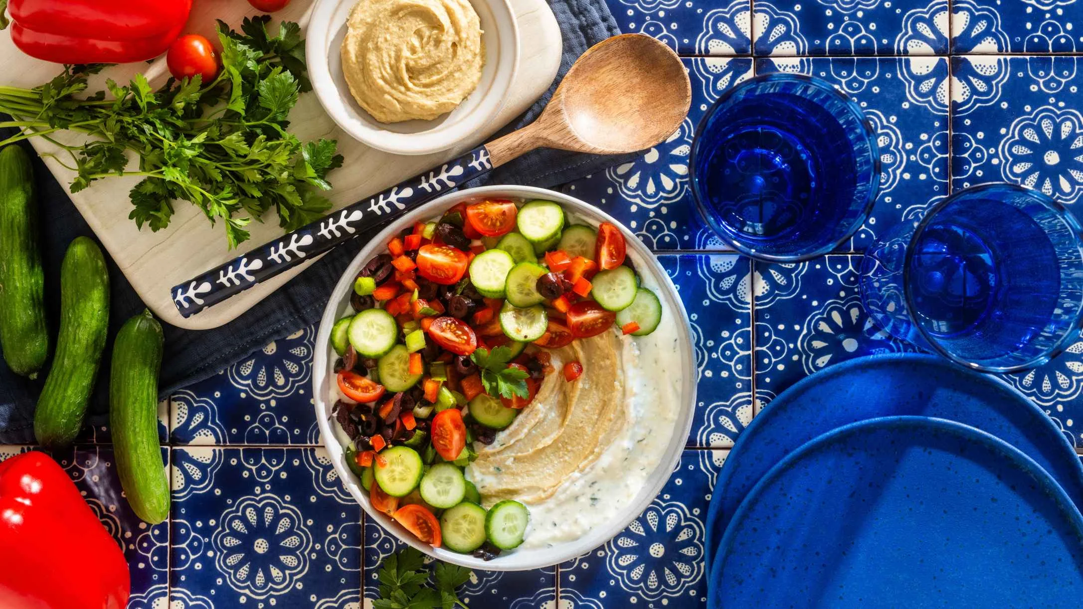 Mediterranean bowl with hummus, fresh vegetables, and salad on blue patterned tablecloth with blue glassware.