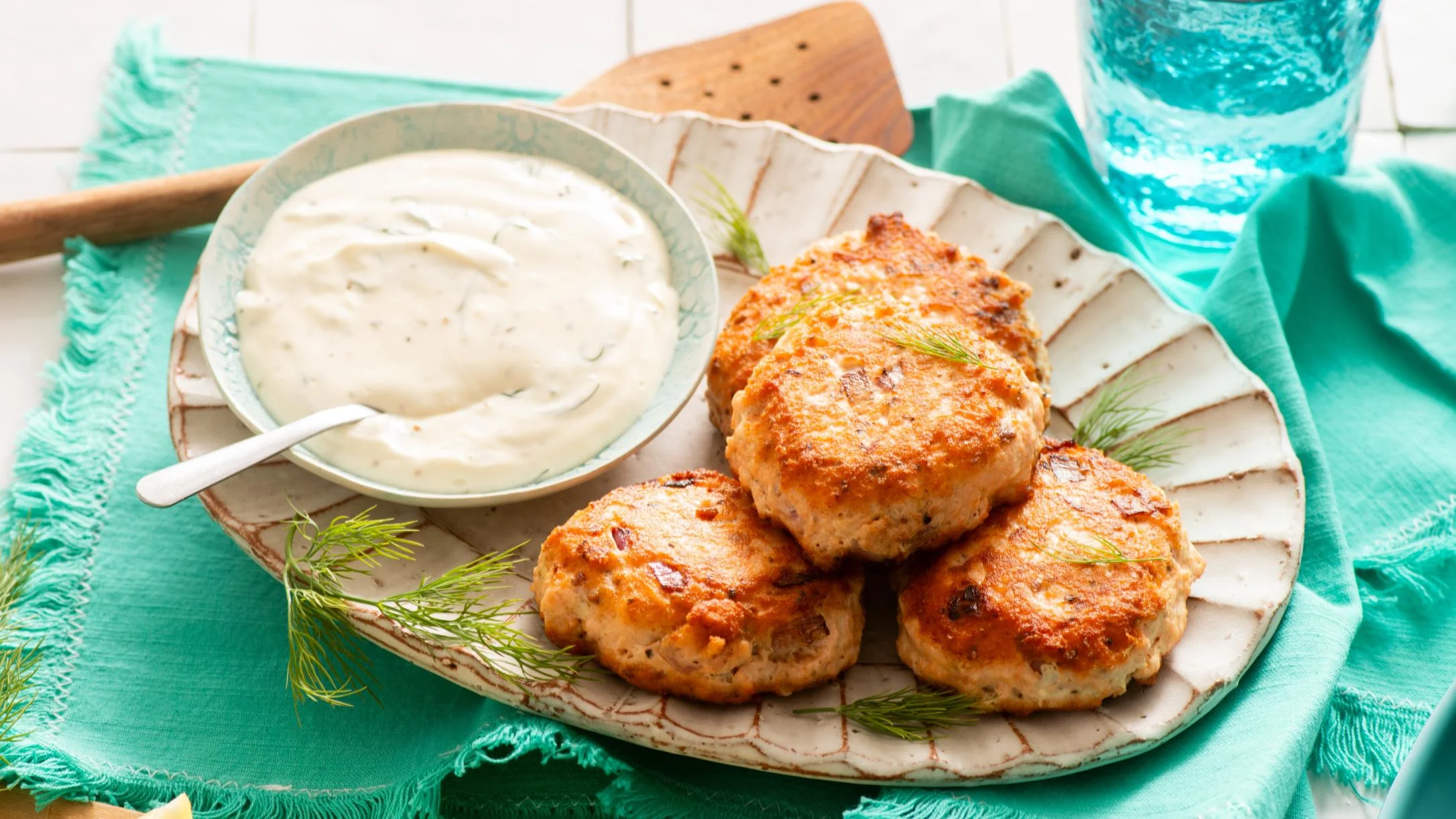 Golden-brown fish cakes garnished with fresh dill on a rustic plate, served with a creamy white dipping sauce on a teal napkin.