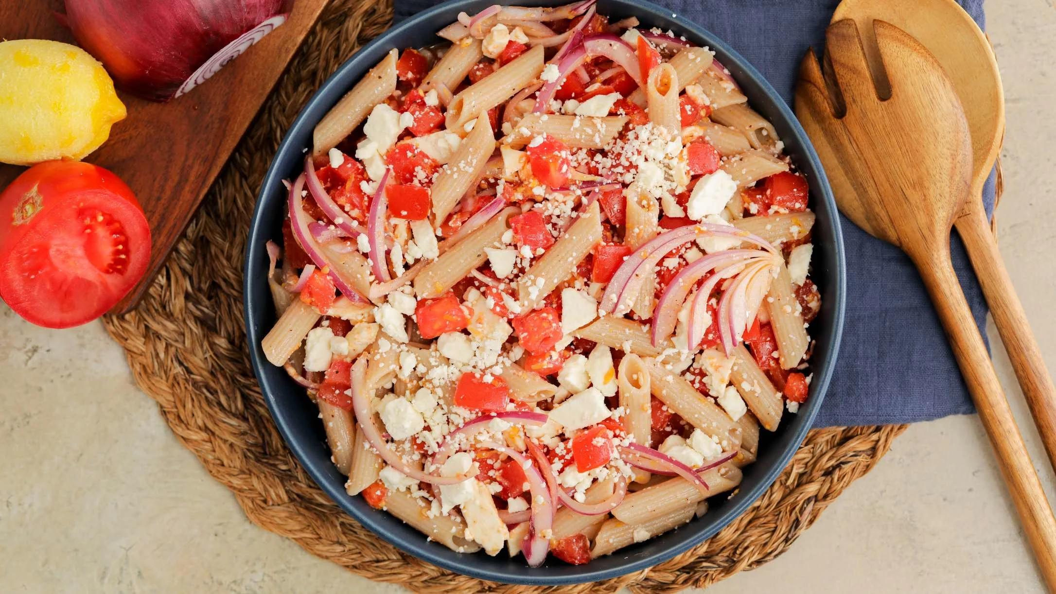 A bowl of penne pasta with tomatoes, red onion, and crumbled feta cheese, beside wooden spoons and fresh tomato and lemon.