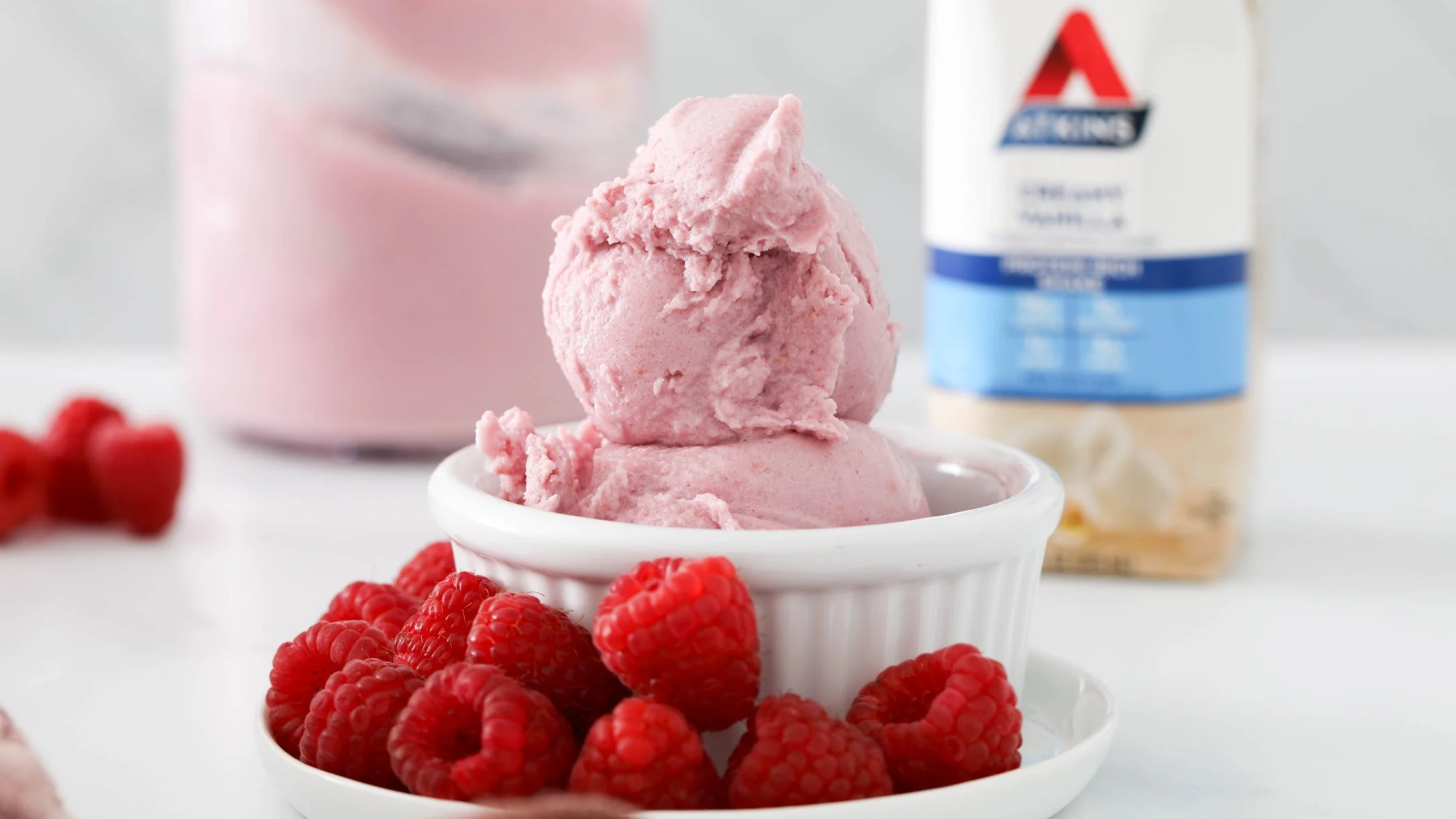 Pink raspberry ice cream scoops in a white ramekin, surrounded by fresh raspberries, with an Atkins shake bottle in the background.
