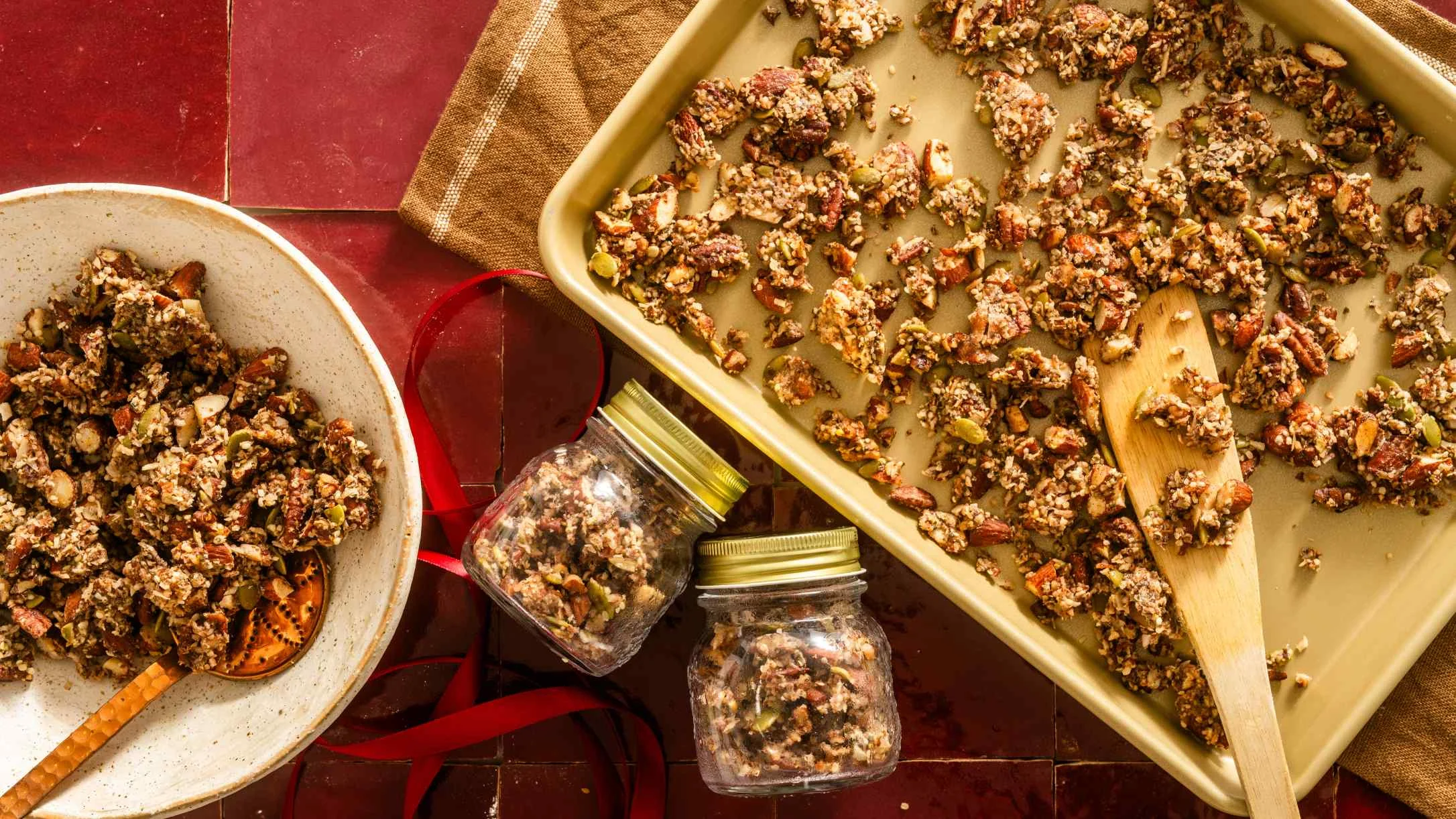 Homemade granola on baking sheet with wooden spoon, bowl of served portion, and small storage jars on red surface.