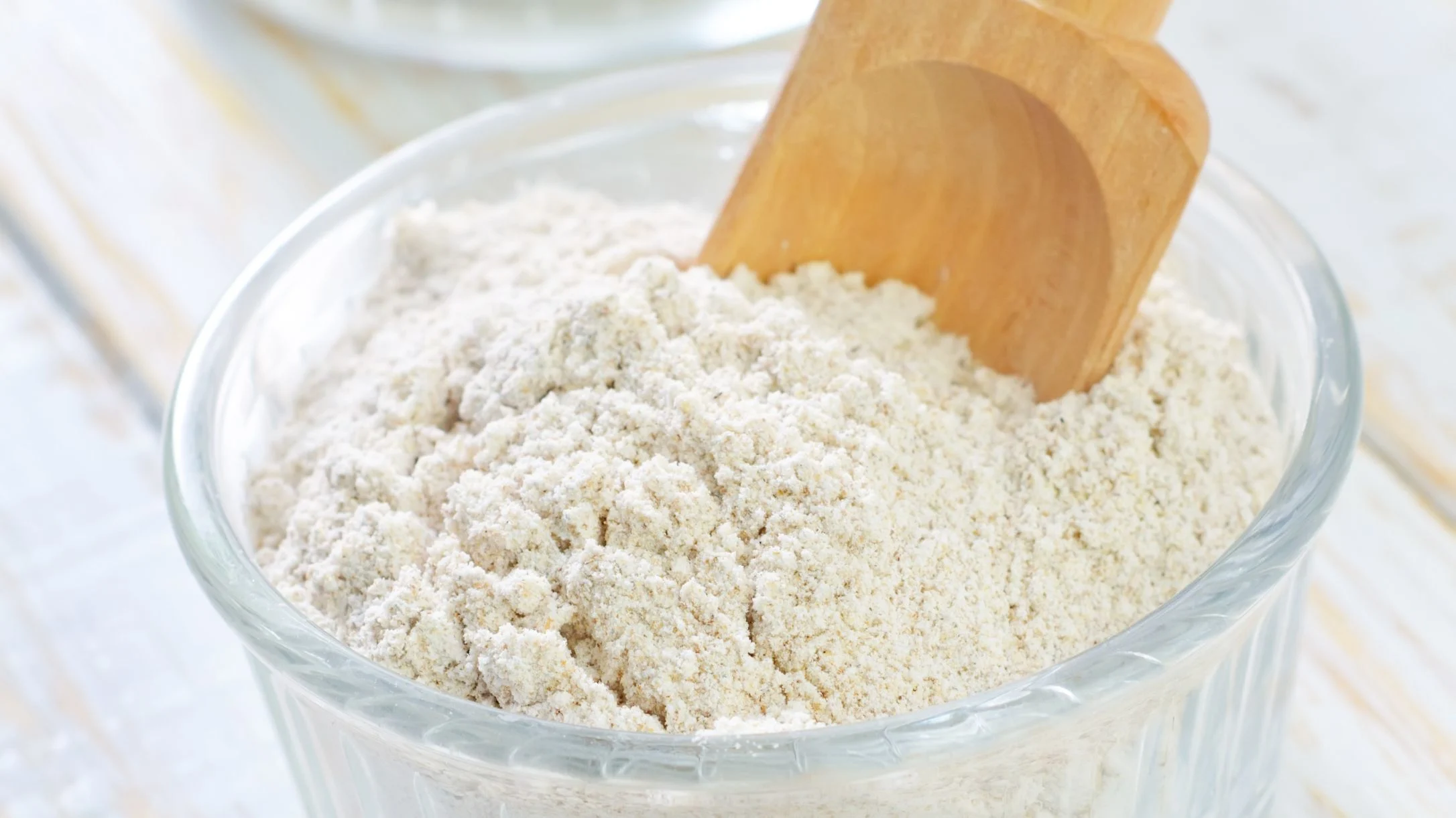 A glass bowl filled with white flour, with a wooden scoop resting inside it on a light wooden surface.