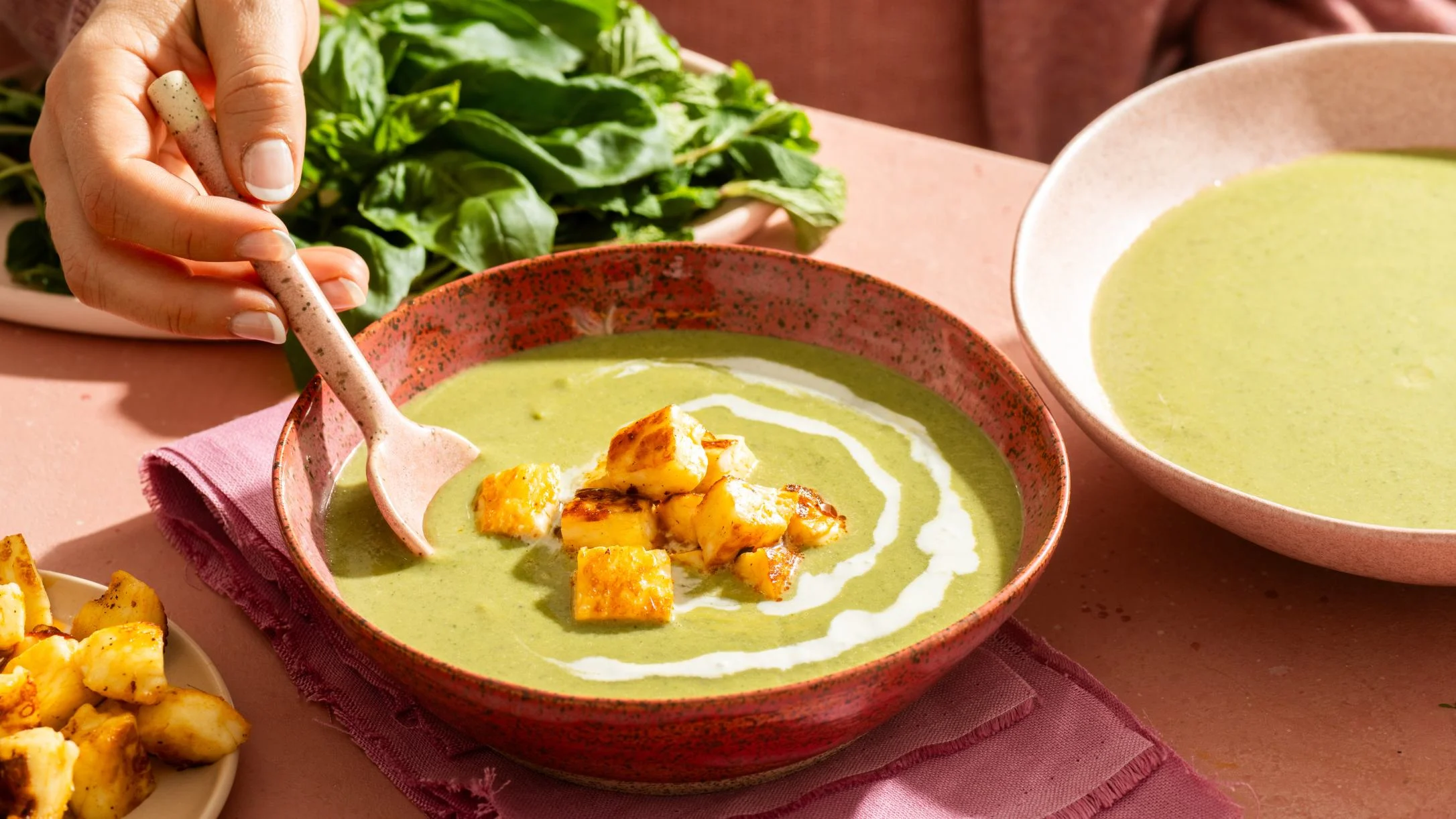 Hand stirring green soup with roasted cheese cubes in a red speckled bowl, fresh herbs nearby.