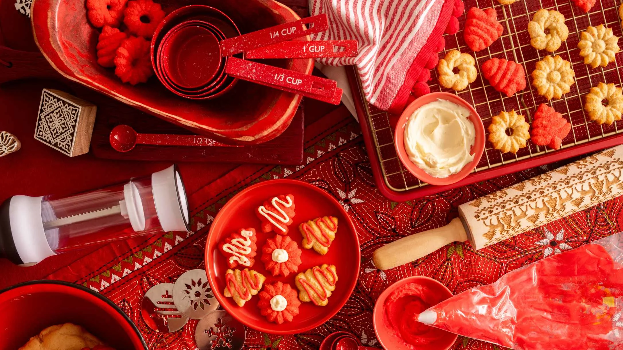 Red holiday baking scene with measuring cups, decorated cookies, frosting, and a wooden rolling pin on a festive tablecloth.