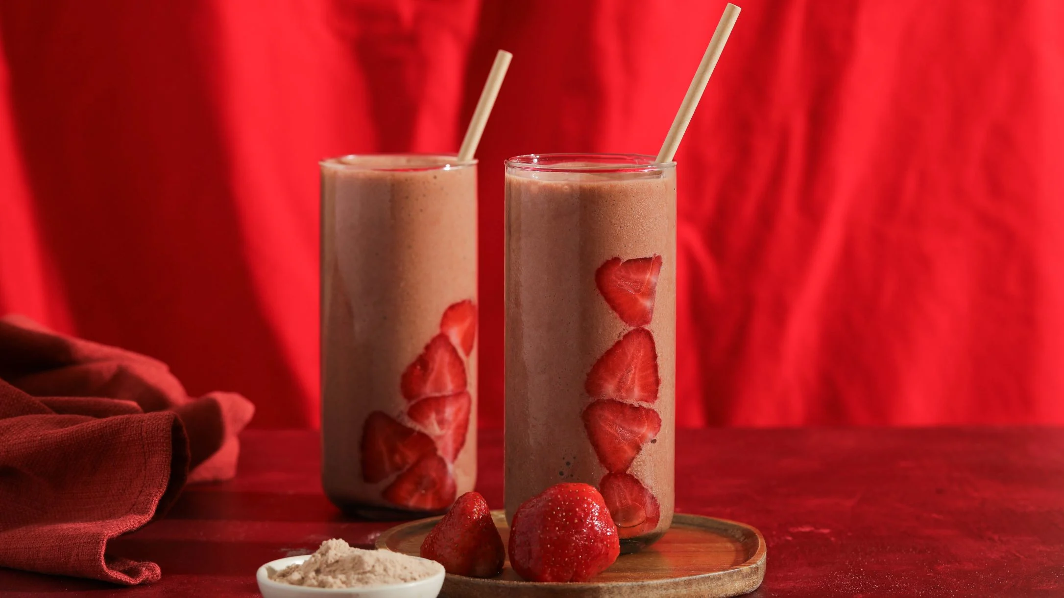 Two tall glasses of strawberry smoothie with paper straws and fresh strawberry slices, on a wooden tray against a red backdrop.