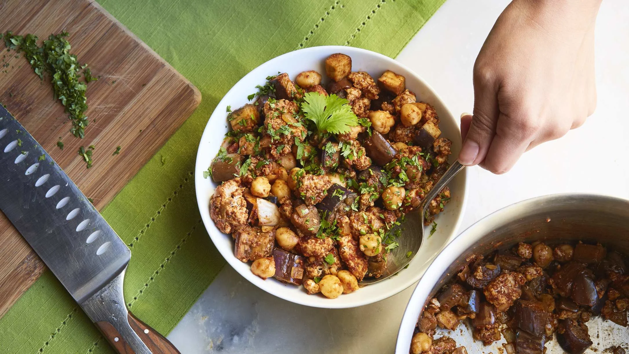 A hand holds a spoon in a white bowl of ground meat and chickpea dish topped with fresh cilantro, beside a cutting board.