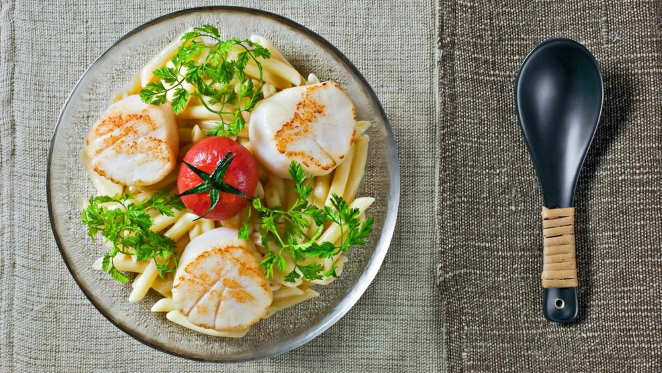 Seared scallops on pasta with fresh herbs and a tomato on a glass plate, beside a black spoon on a linen surface.