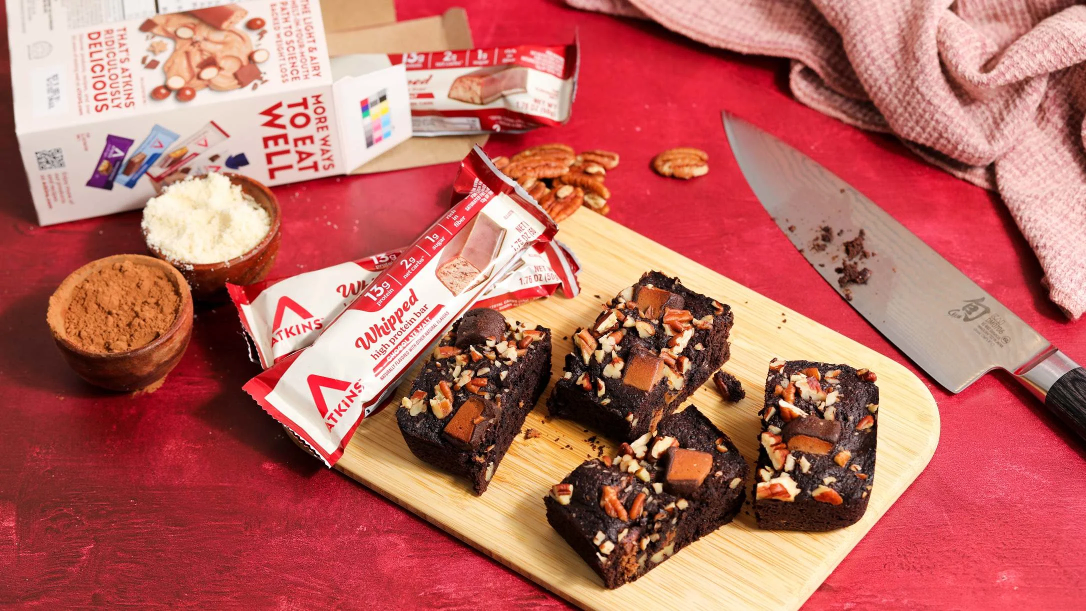 Chocolate pecan brownies on a cutting board with Atkins Whipped protein bars, cocoa powder, and product boxes on a red surface.