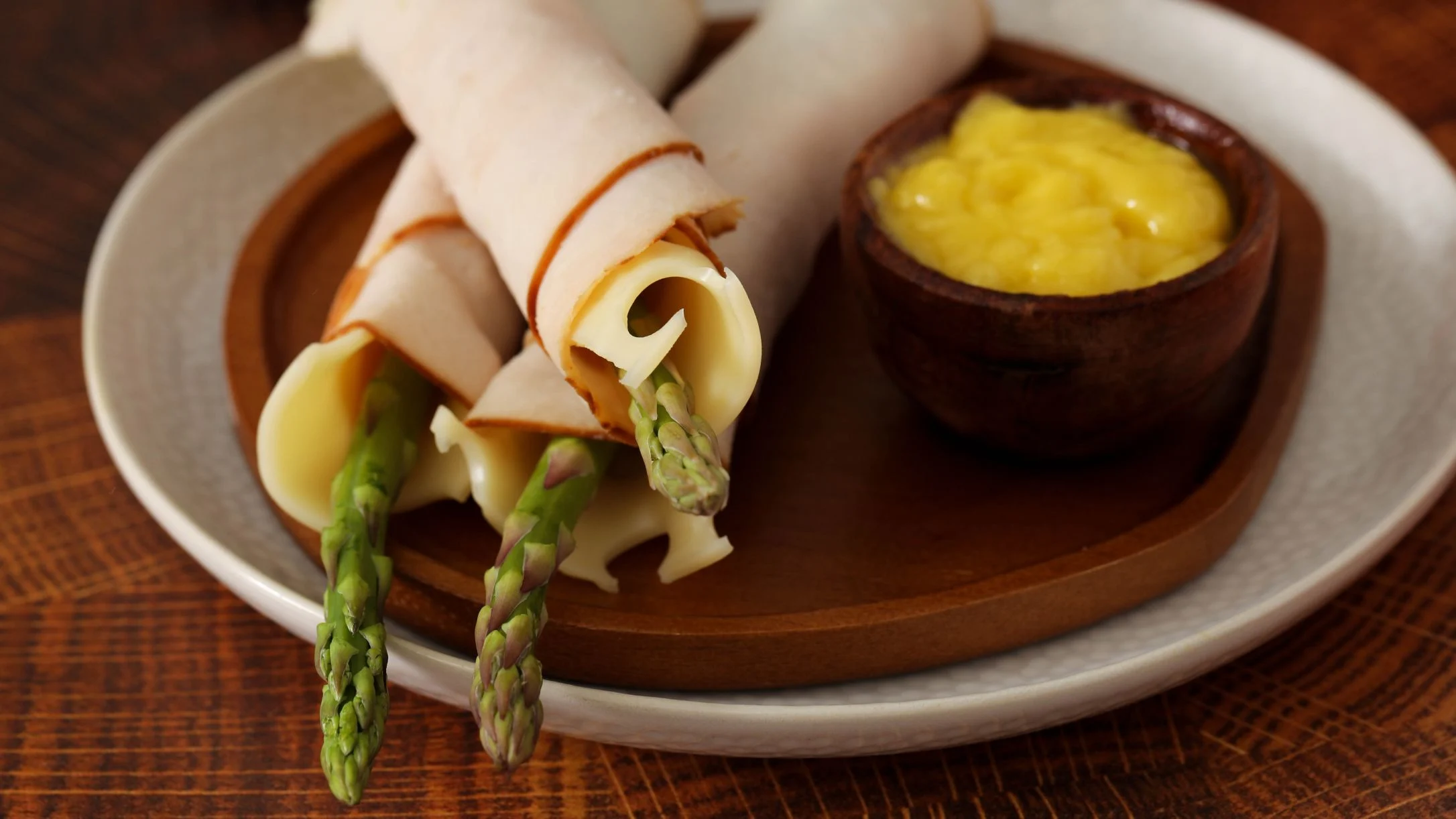 Turkey and cheese rolls wrapped around fresh asparagus spears on a plate, served with a small bowl of yellow aioli.