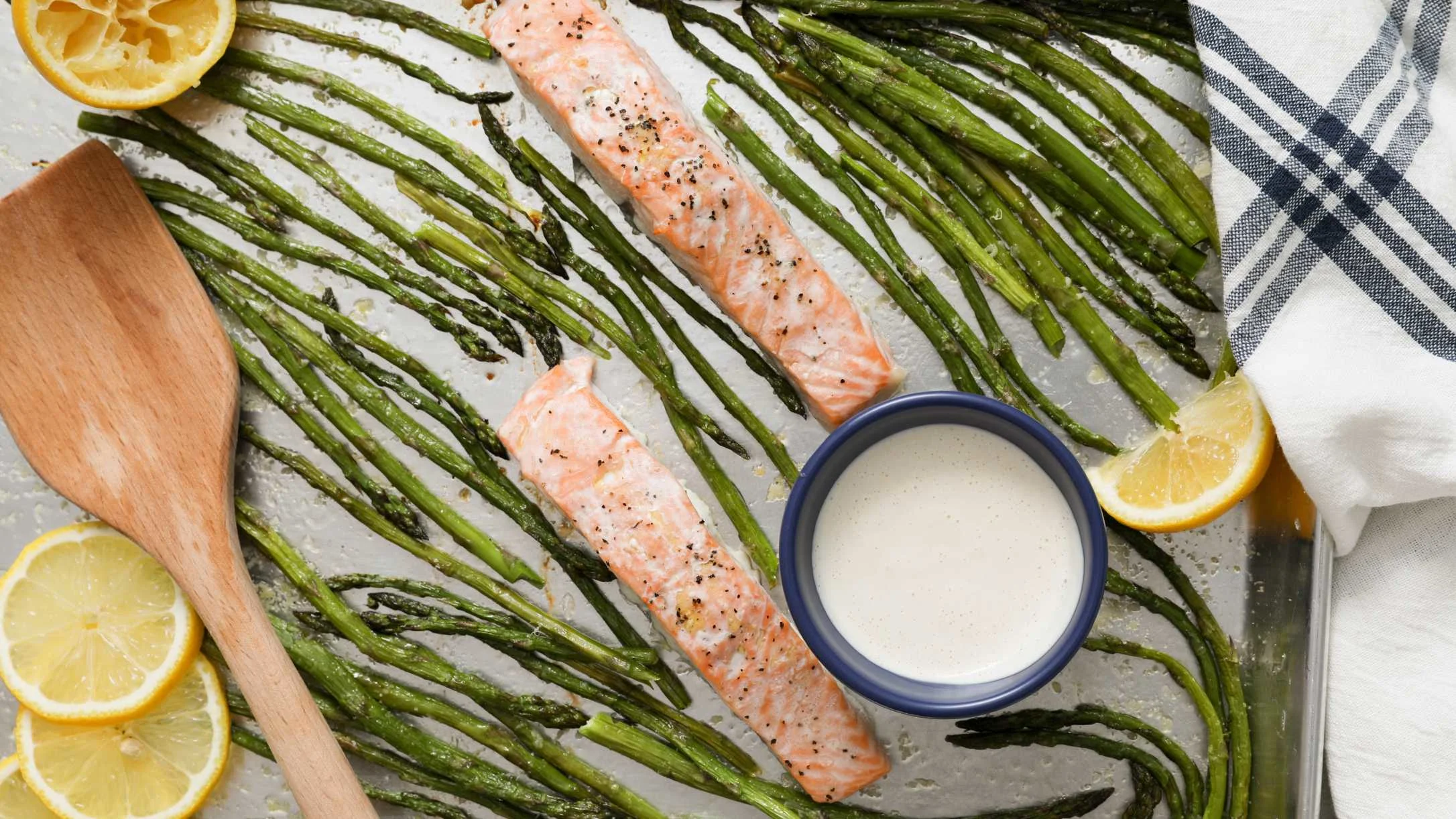 Two roasted salmon fillets and asparagus on a sheet pan with lemon slices, a creamy dipping sauce, and a wooden spoon.
