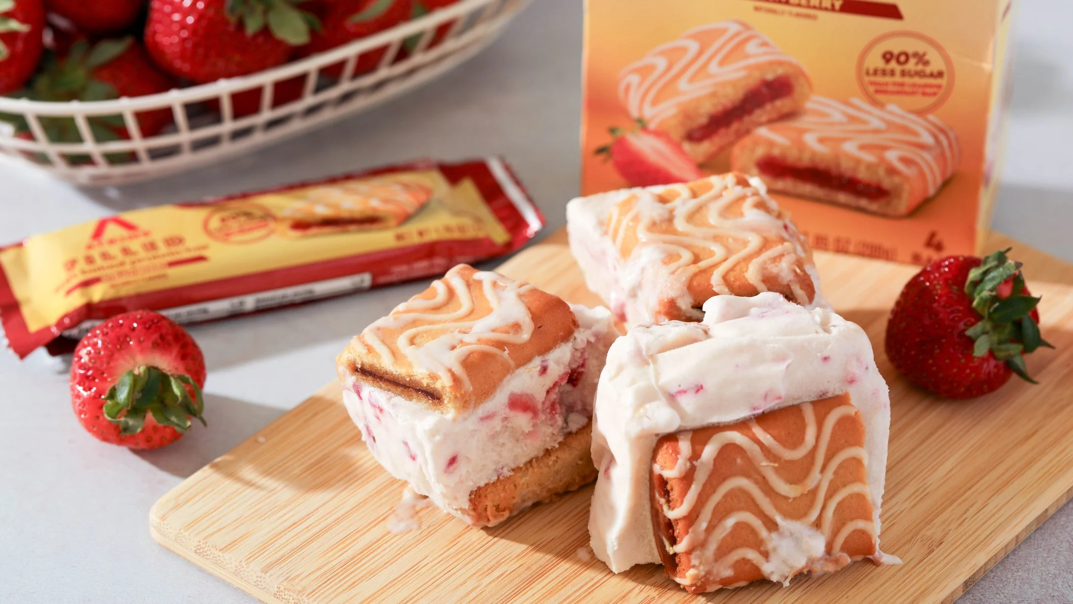 Sliced strawberry cream-filled snack bars on a wooden board, with fresh strawberries, product packaging, and a wrapper nearby.