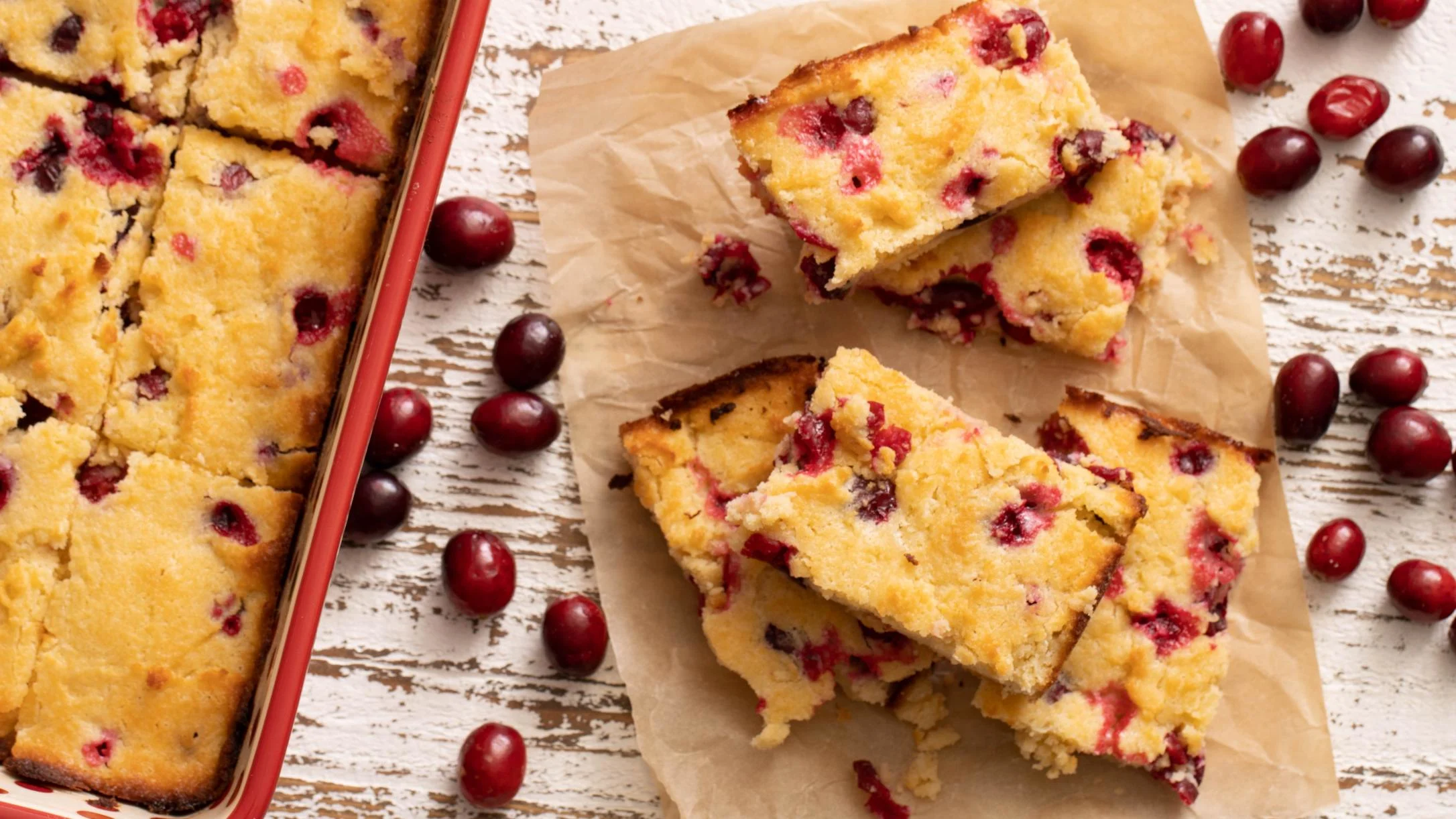 Sliced cranberry cake bars on parchment paper beside a red baking pan, with fresh cranberries scattered on a rustic white surface.