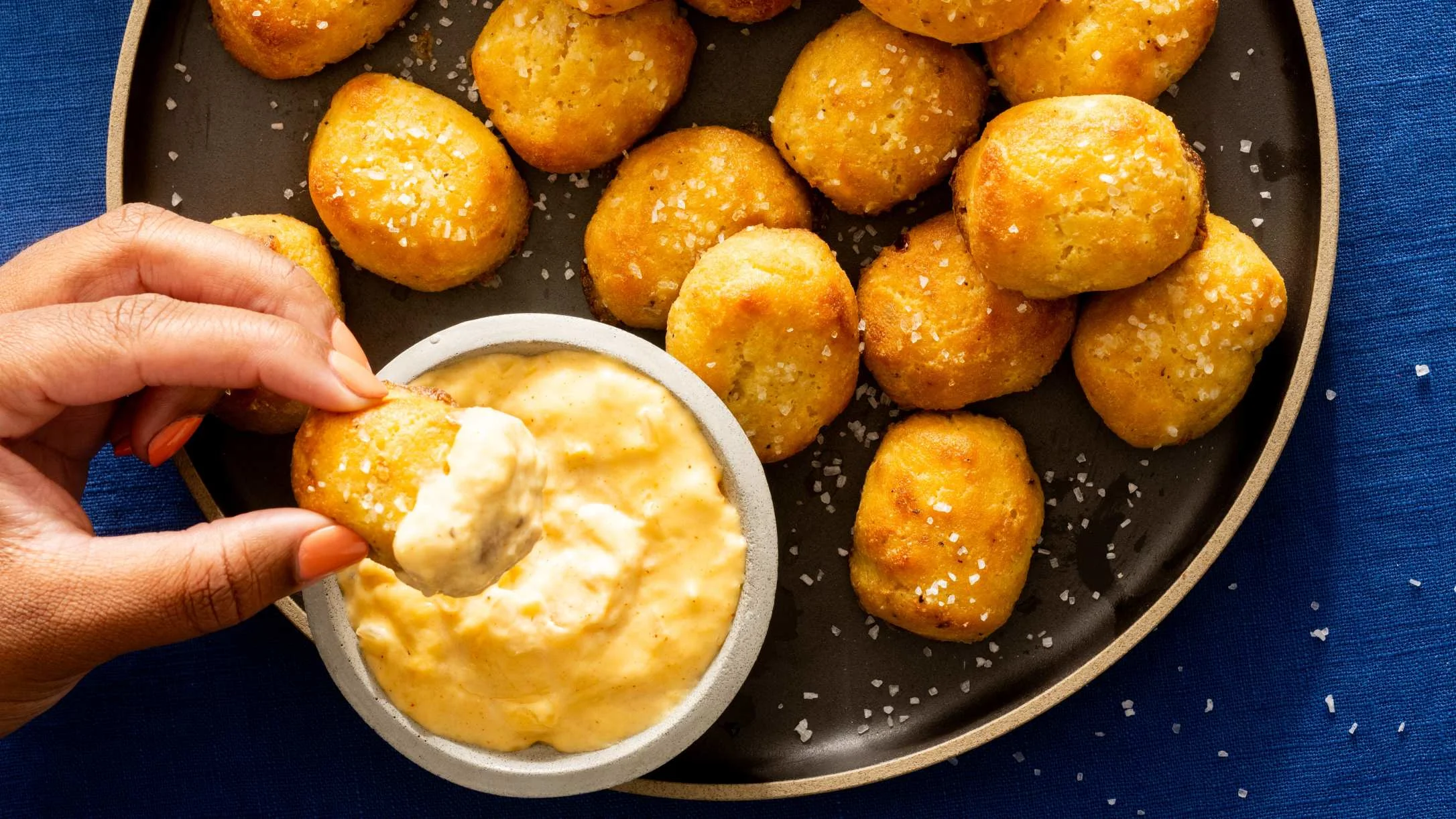 A hand dips a golden, salted pretzel bite into a creamy dipping sauce, with more bites on a dark plate nearby.