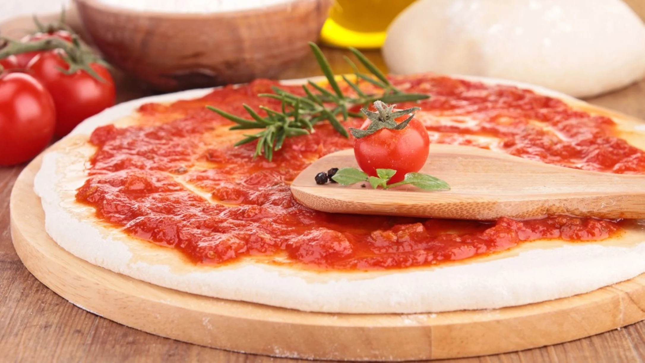 Pizza dough topped with red tomato sauce on a wooden board, with a cherry tomato, rosemary, and a wooden spatula on top.
