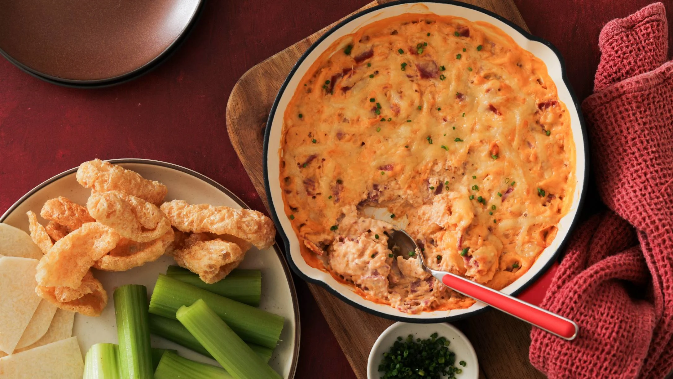 Baked reuben dip in a skillet with a spoon, served alongside pork rinds, celery sticks, and jicama slices on a plate.