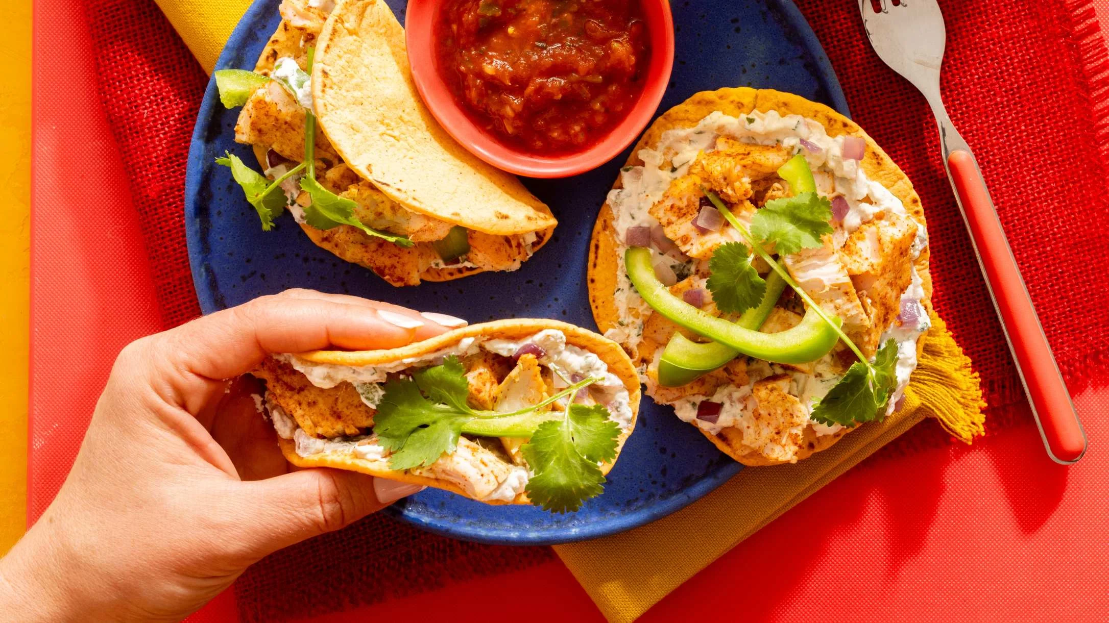 A hand holds a turkey taco topped with cream sauce, peppers, and cilantro on a blue plate with salsa.