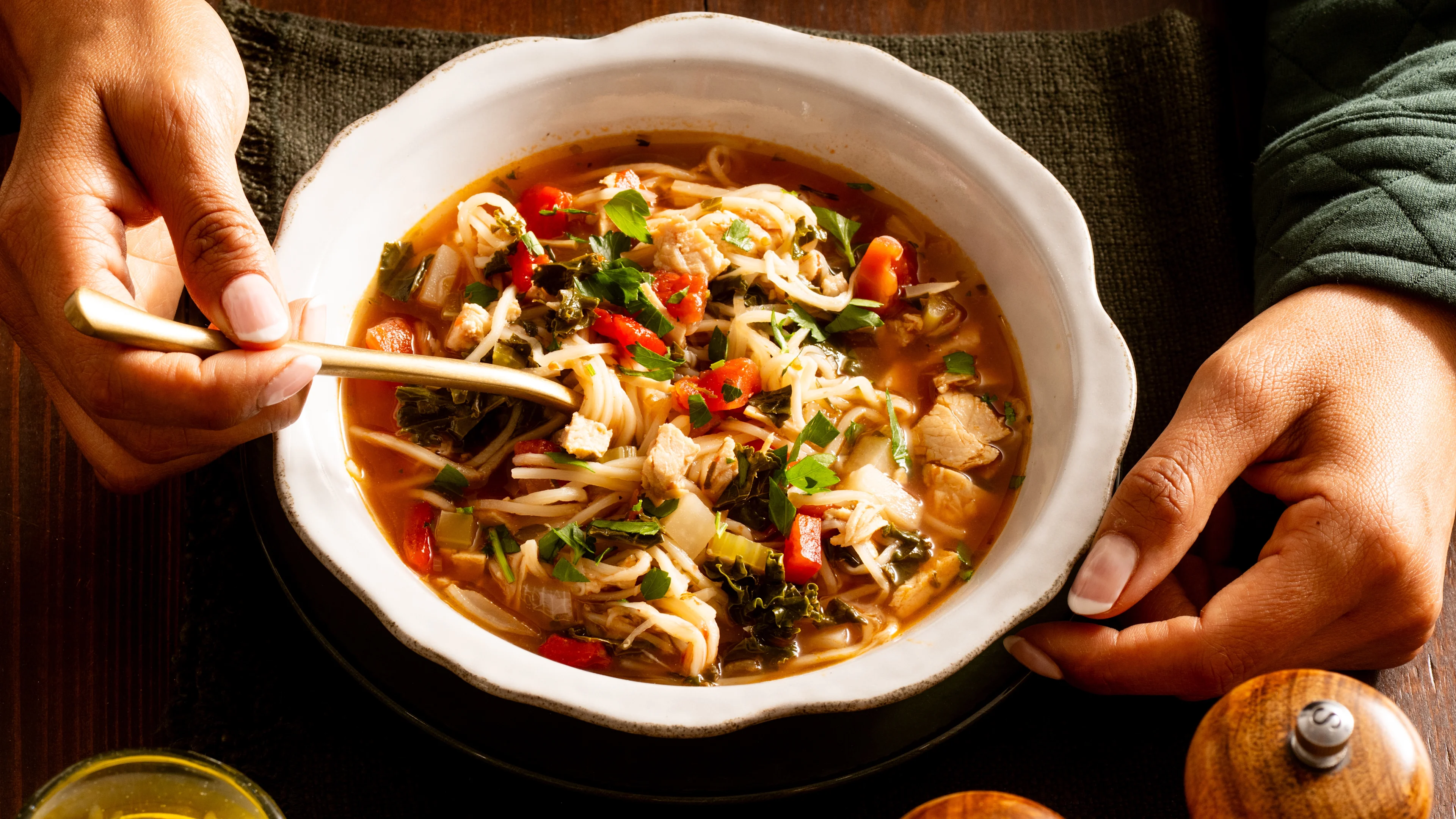 Hands holding a white bowl of hearty turkey noodle soup with kale, red peppers, and herbs on a dark wooden table.