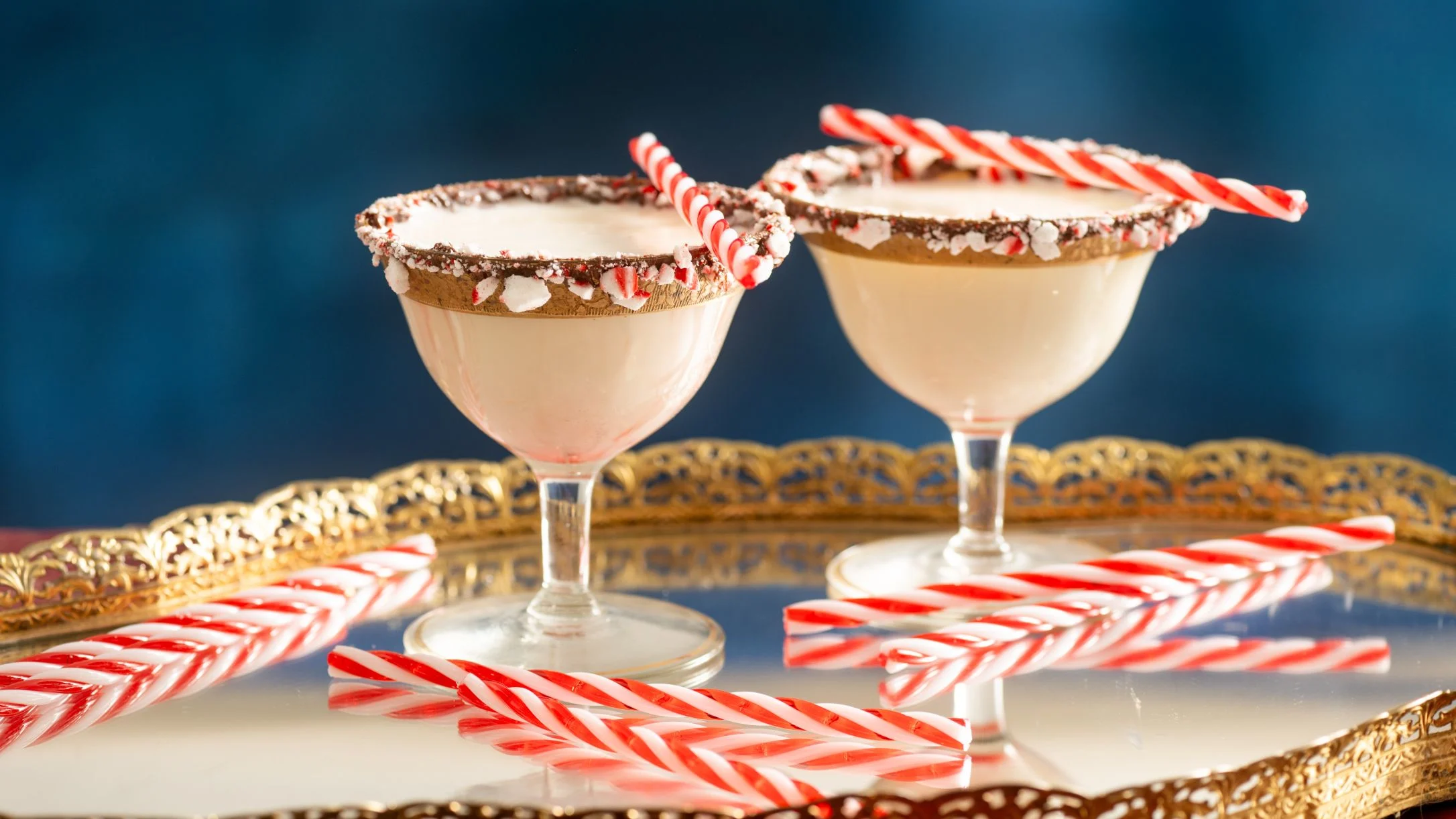 Two festive cocktails with crushed candy cane rims and peppermint sticks on a gold tray against a blue background.