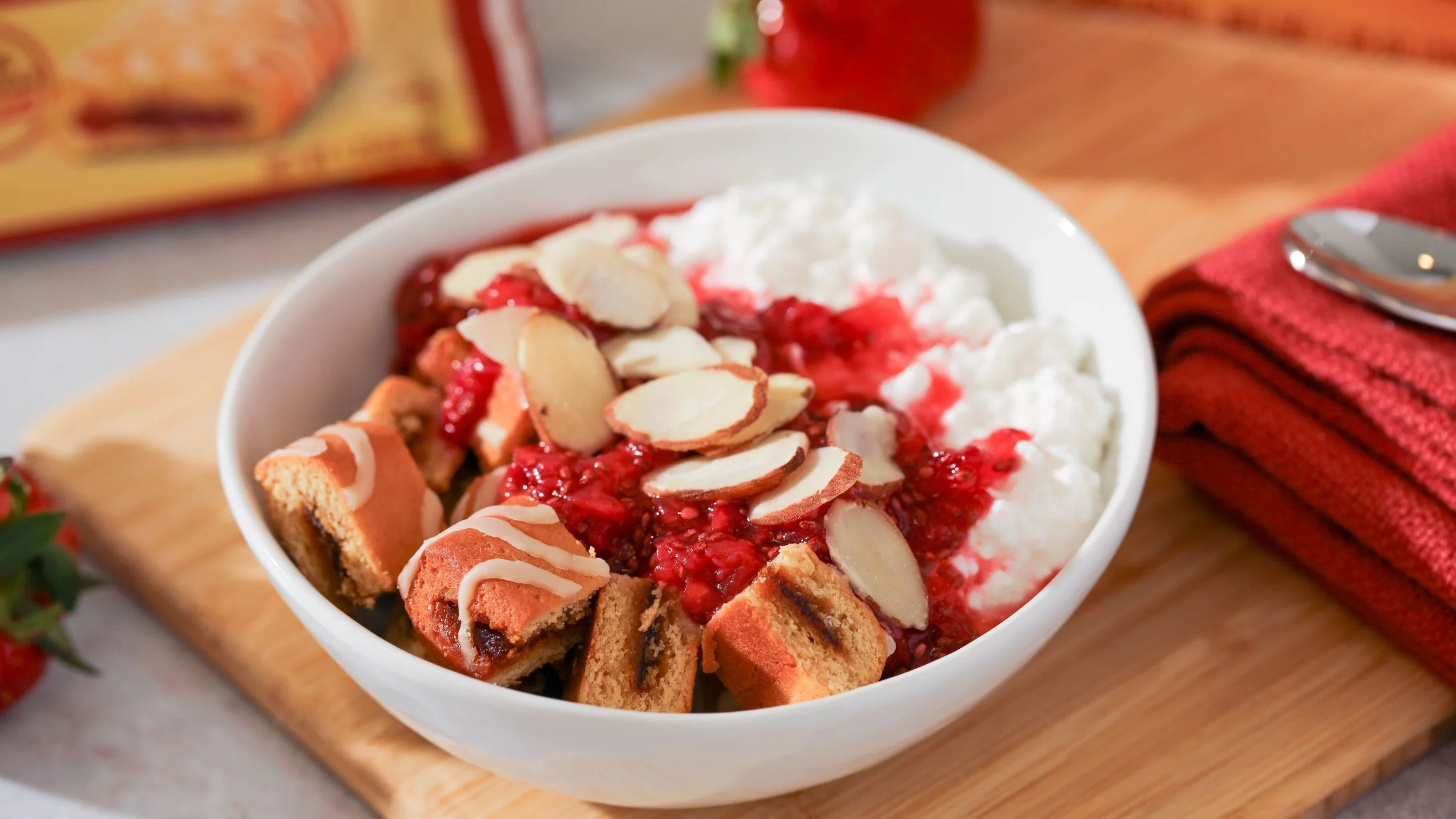 White bowl with Atkins bar pieces, strawberry sauce, sliced almonds, and cottage cheese on a wooden cutting board.