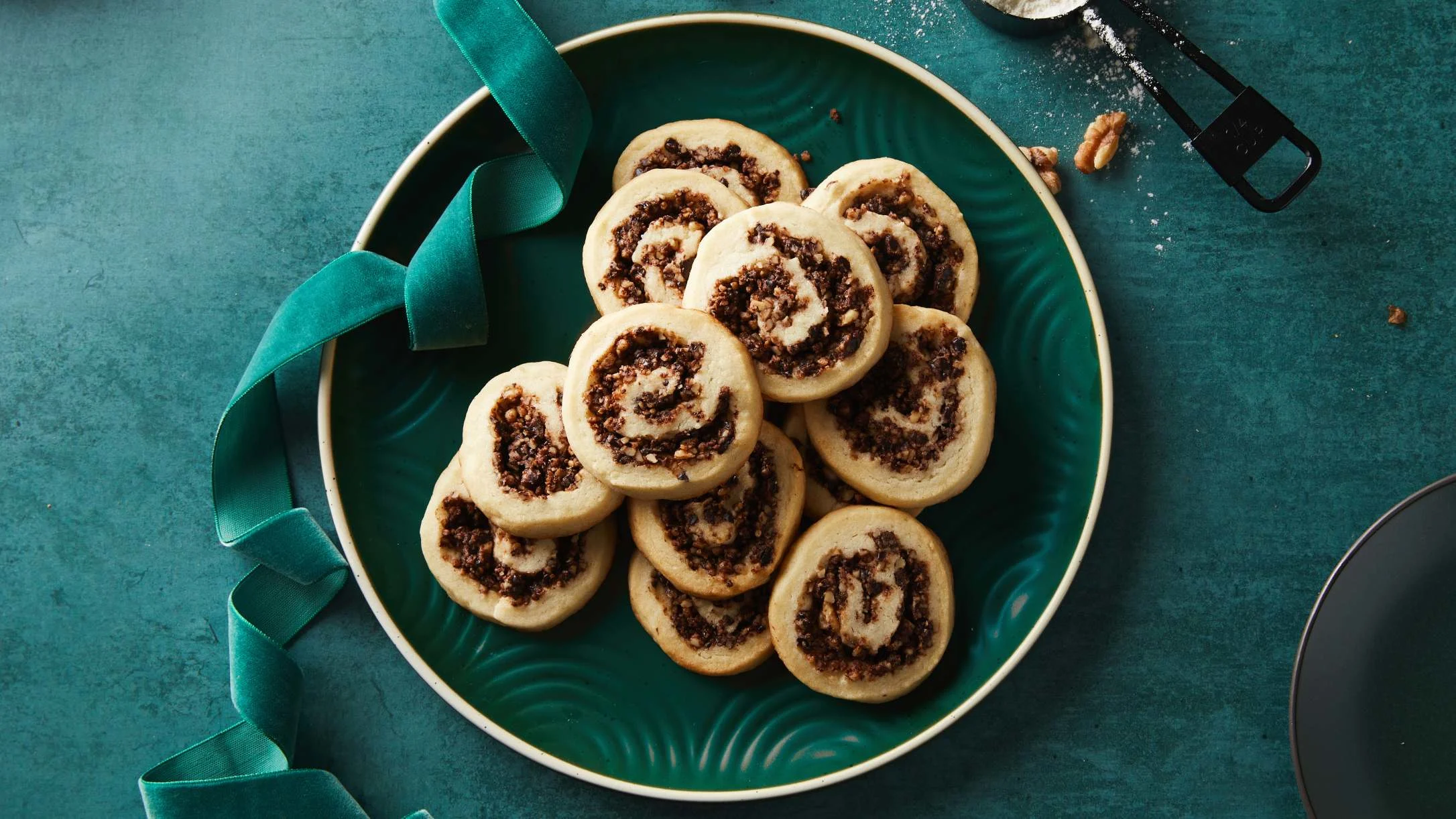 Pinwheel cookies with nut filling arranged on a teal plate with matching ribbon on teal background.
