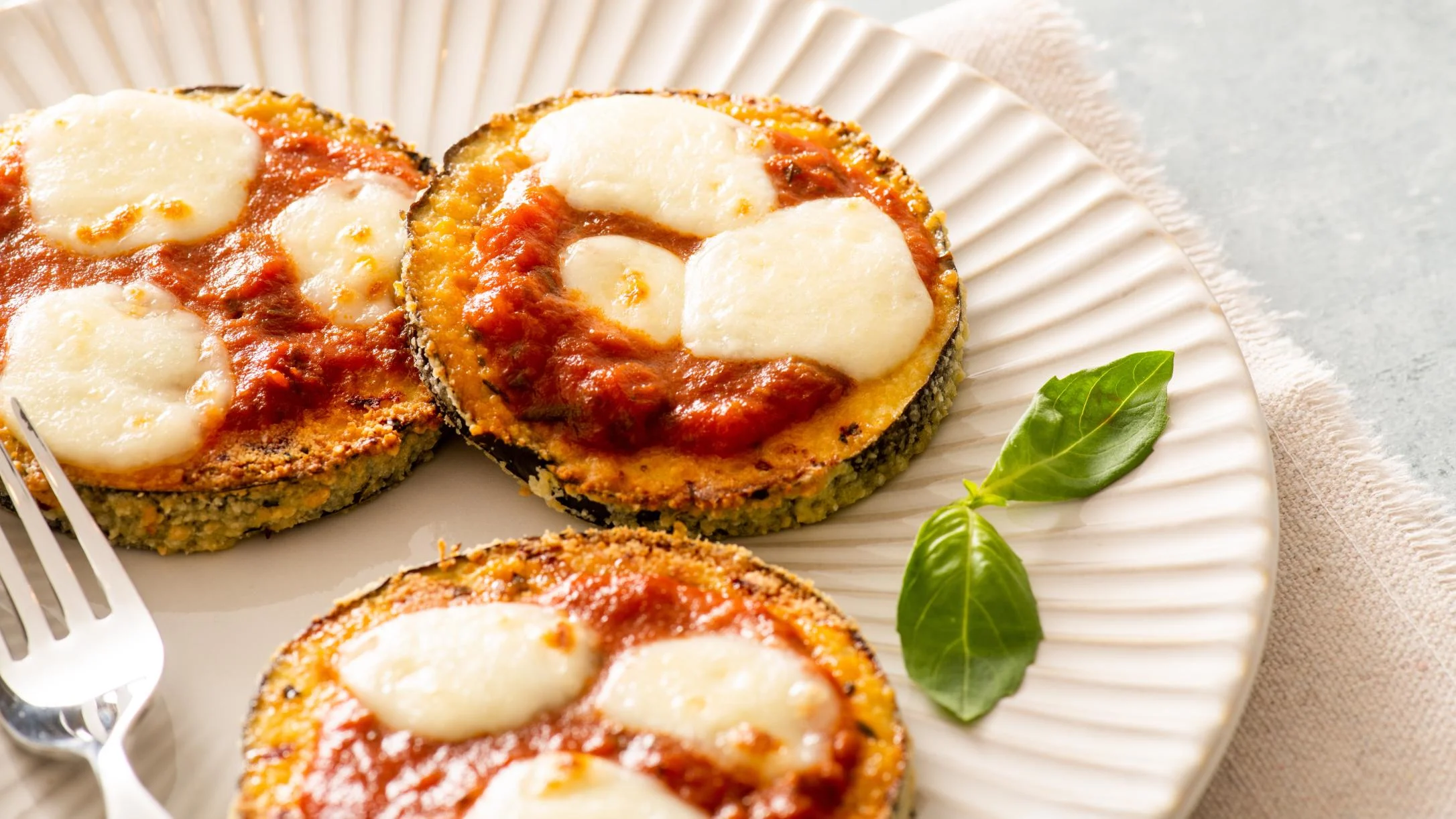 Three breaded eggplant rounds topped with tomato sauce and melted mozzarella on a white plate with fresh basil.
