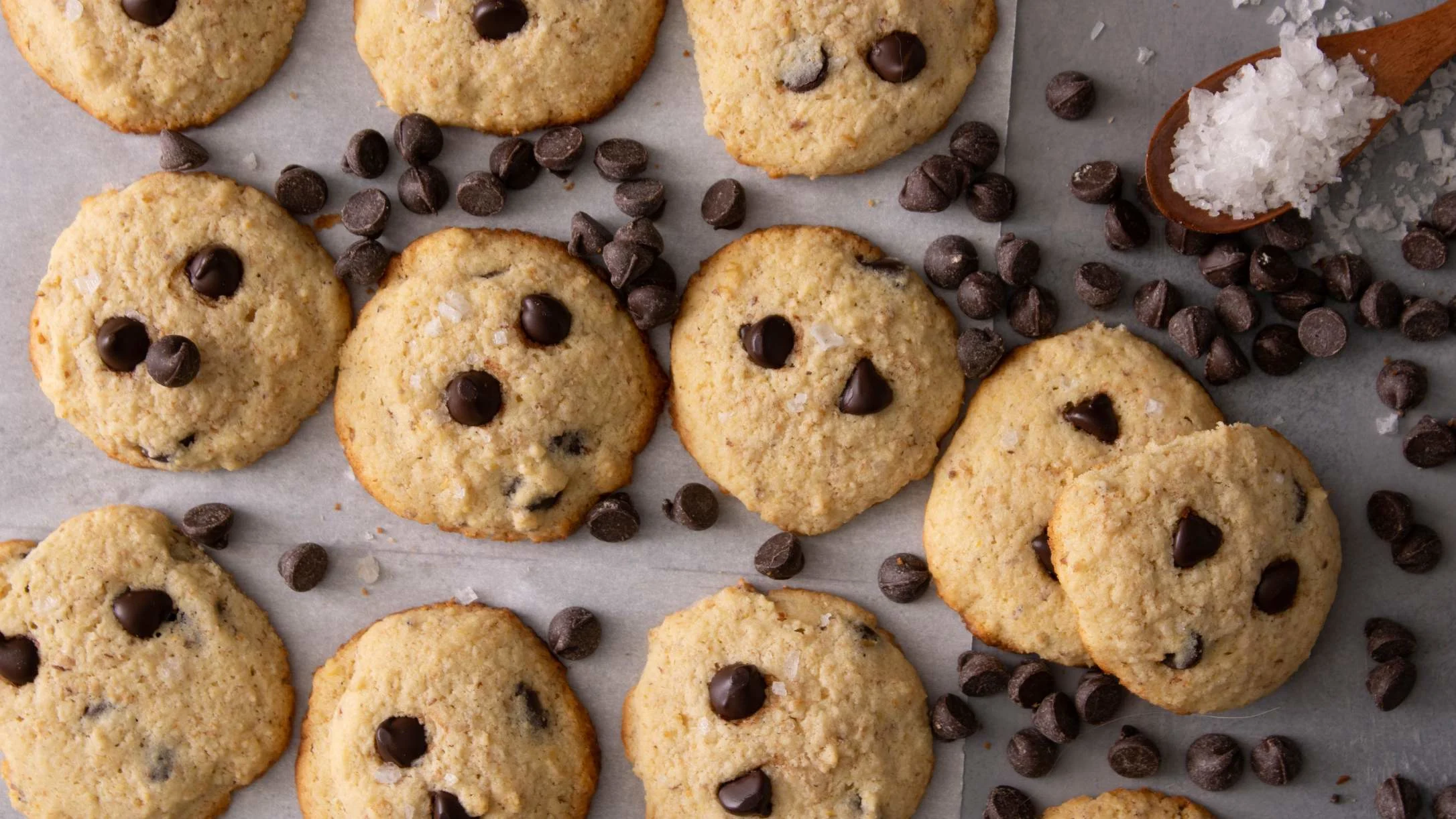 Freshly baked chocolate chip cookies on parchment paper with scattered chocolate chips and a wooden spoon of sea salt.
