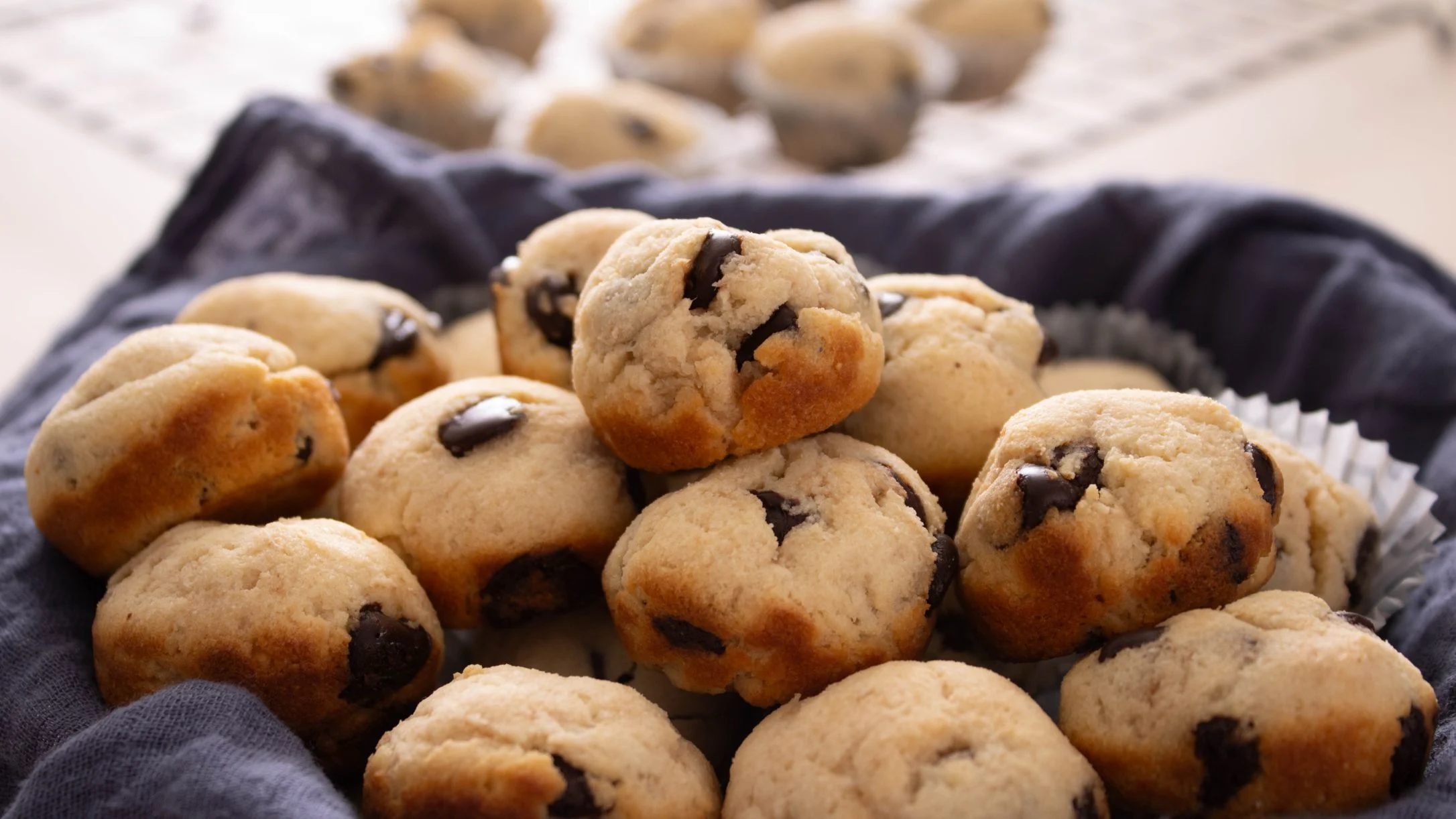 A pile of golden mini chocolate chip muffins stacked in a blue cloth-lined basket, with more muffins blurred in the background.