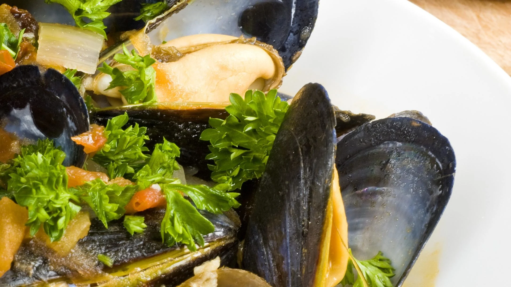 Steamed mussels in their dark shells served on a white plate with fresh parsley, diced tomatoes, and vegetables in broth.