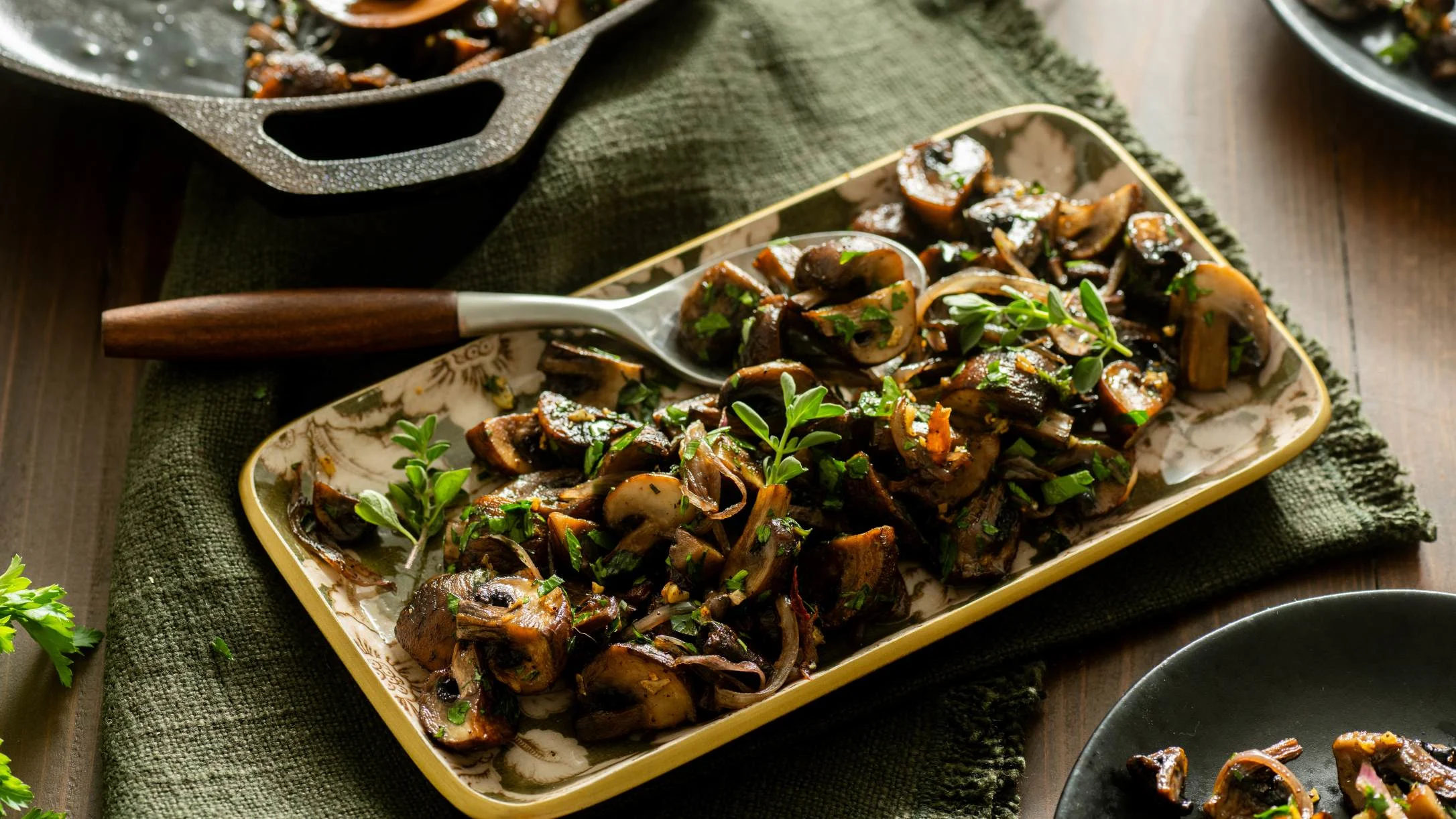 Sautéed mushrooms with caramelized onions and fresh herbs on a decorative platter, with a spoon and skillet nearby.