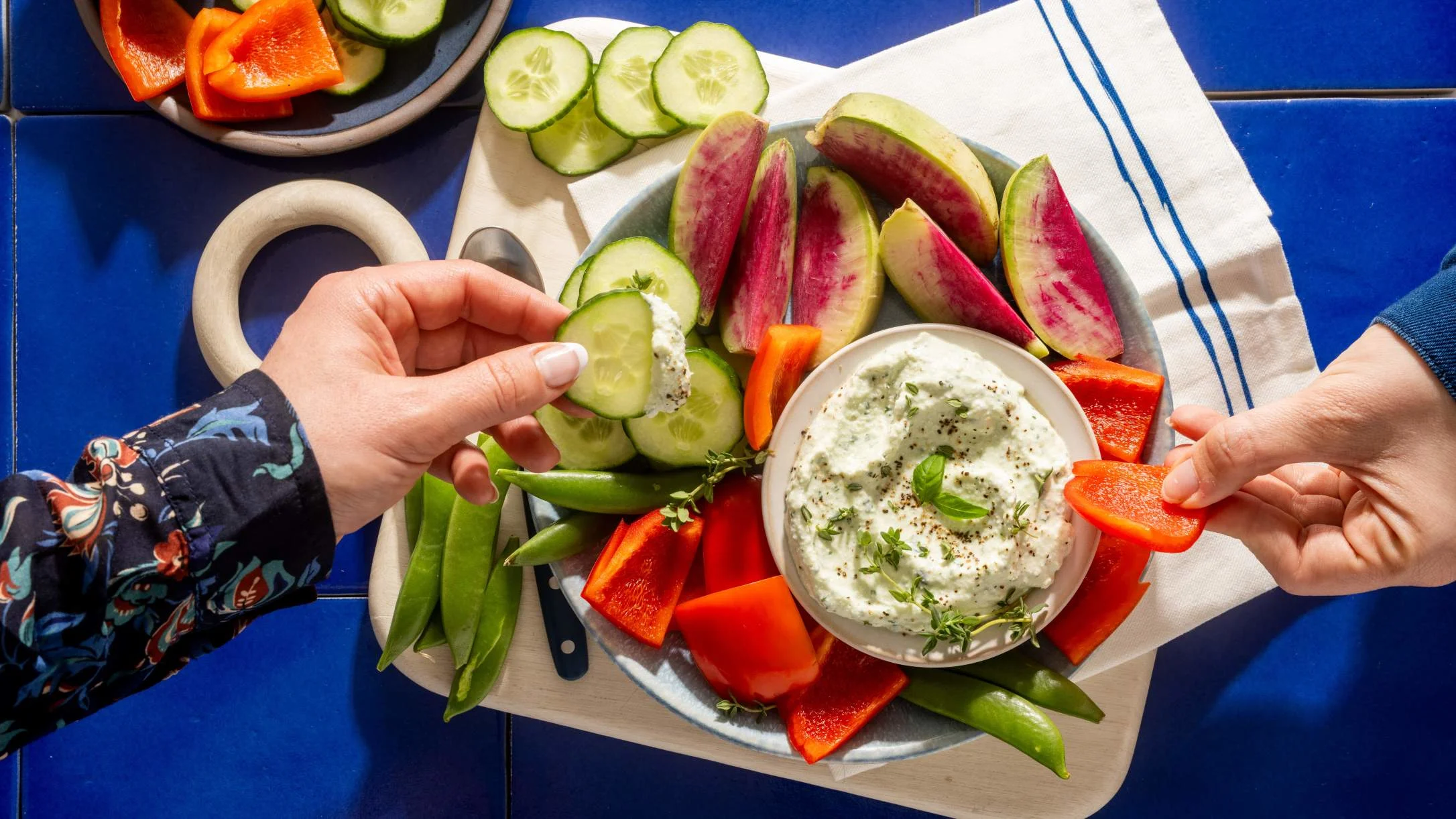 Hands reaching for fresh vegetable platter with herb dip, featuring cucumber slices, radishes, bell peppers and snap peas.