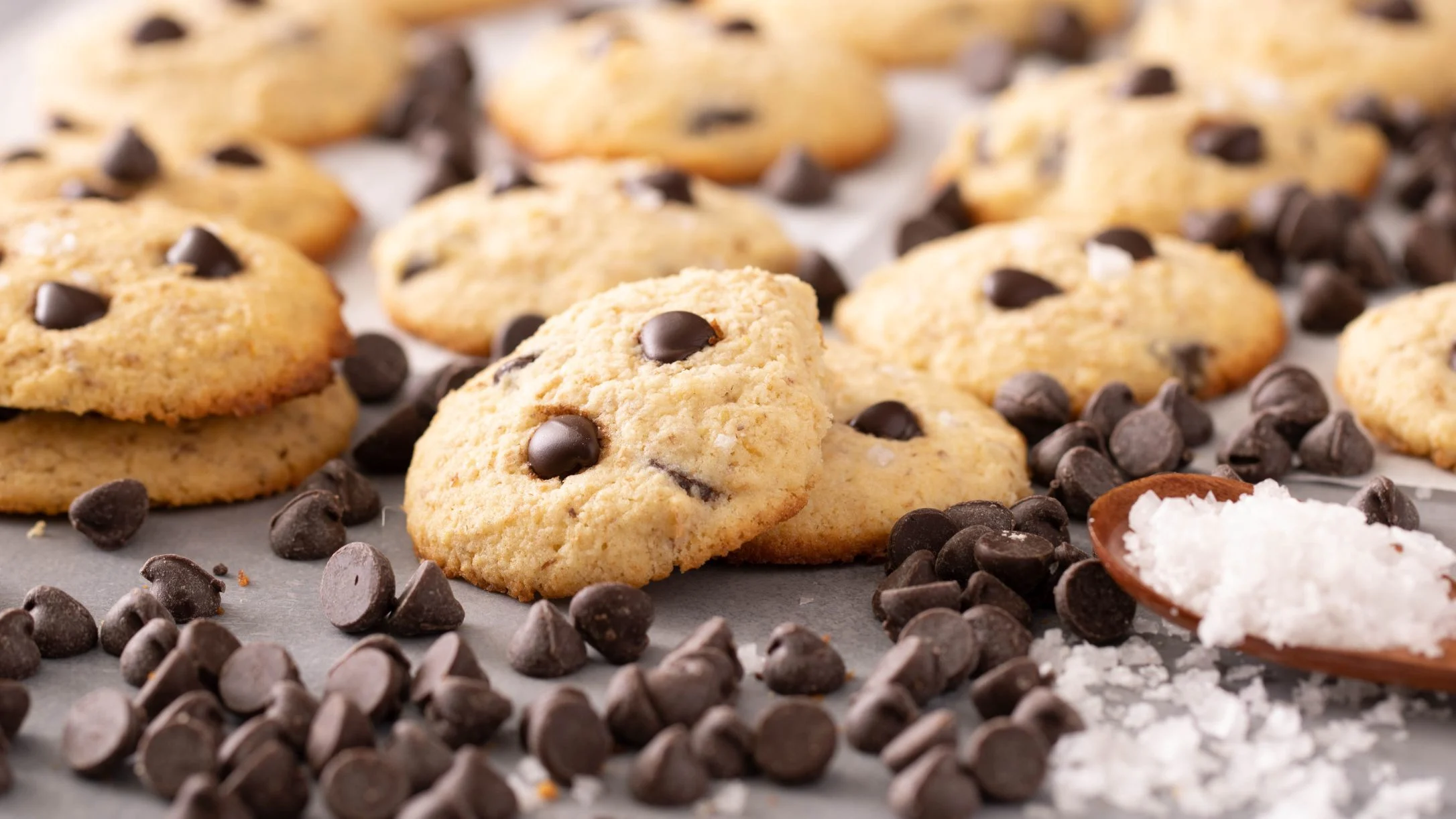 Freshly baked chocolate chip cookies on a gray surface with scattered chocolate chips and a small dish of salt.