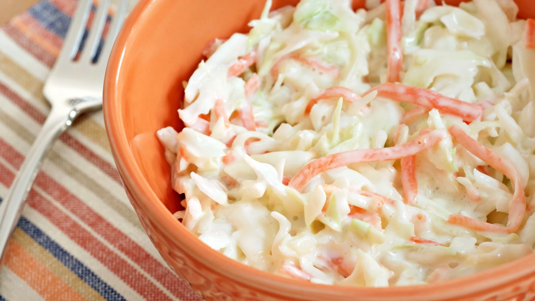Creamy coleslaw with shredded cabbage and carrots in an orange bowl, placed on a striped cloth with a fork nearby.
