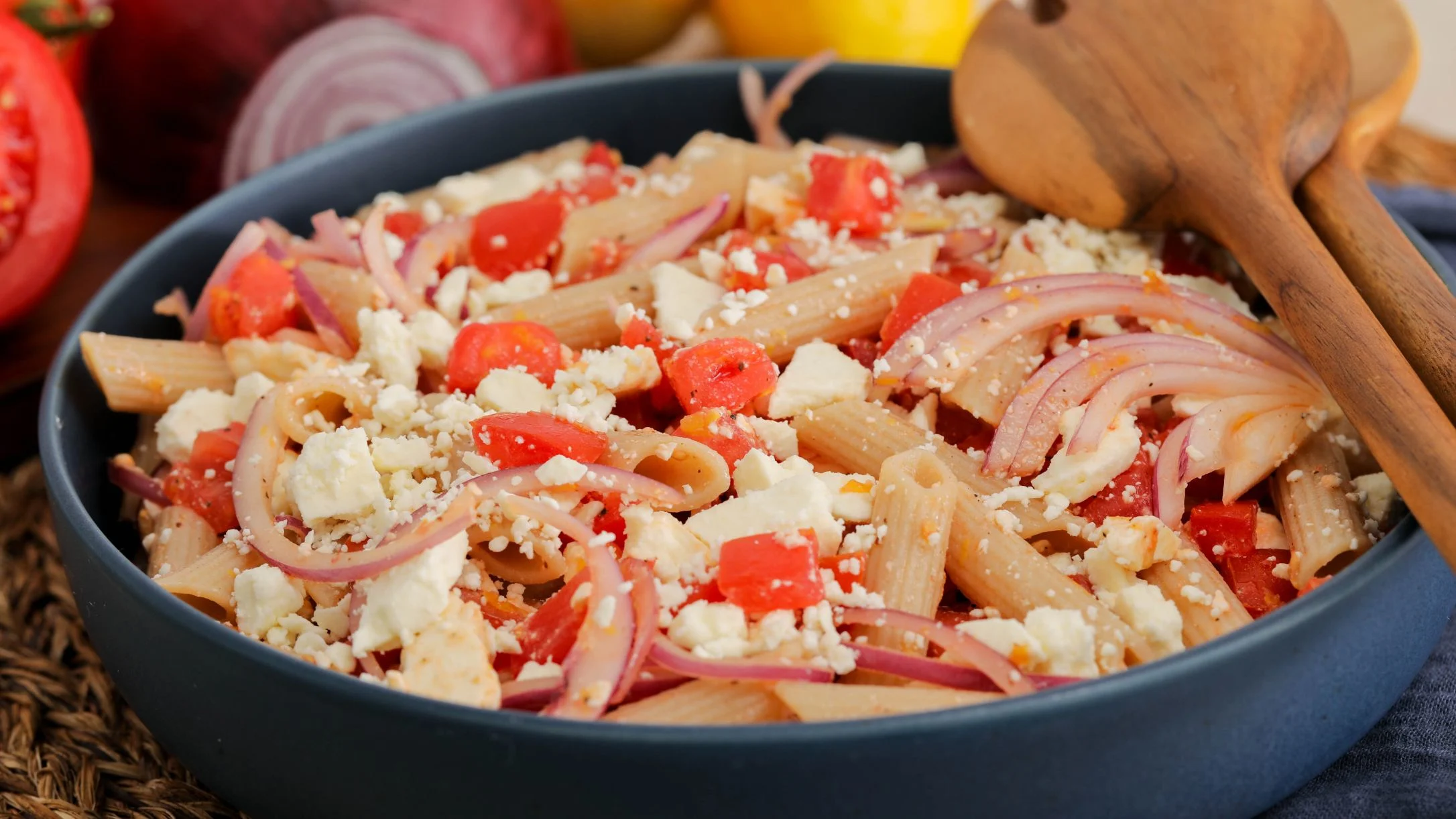 A blue bowl of penne pasta salad with crumbled feta, diced tomatoes, and red onion, served with wooden spoons.