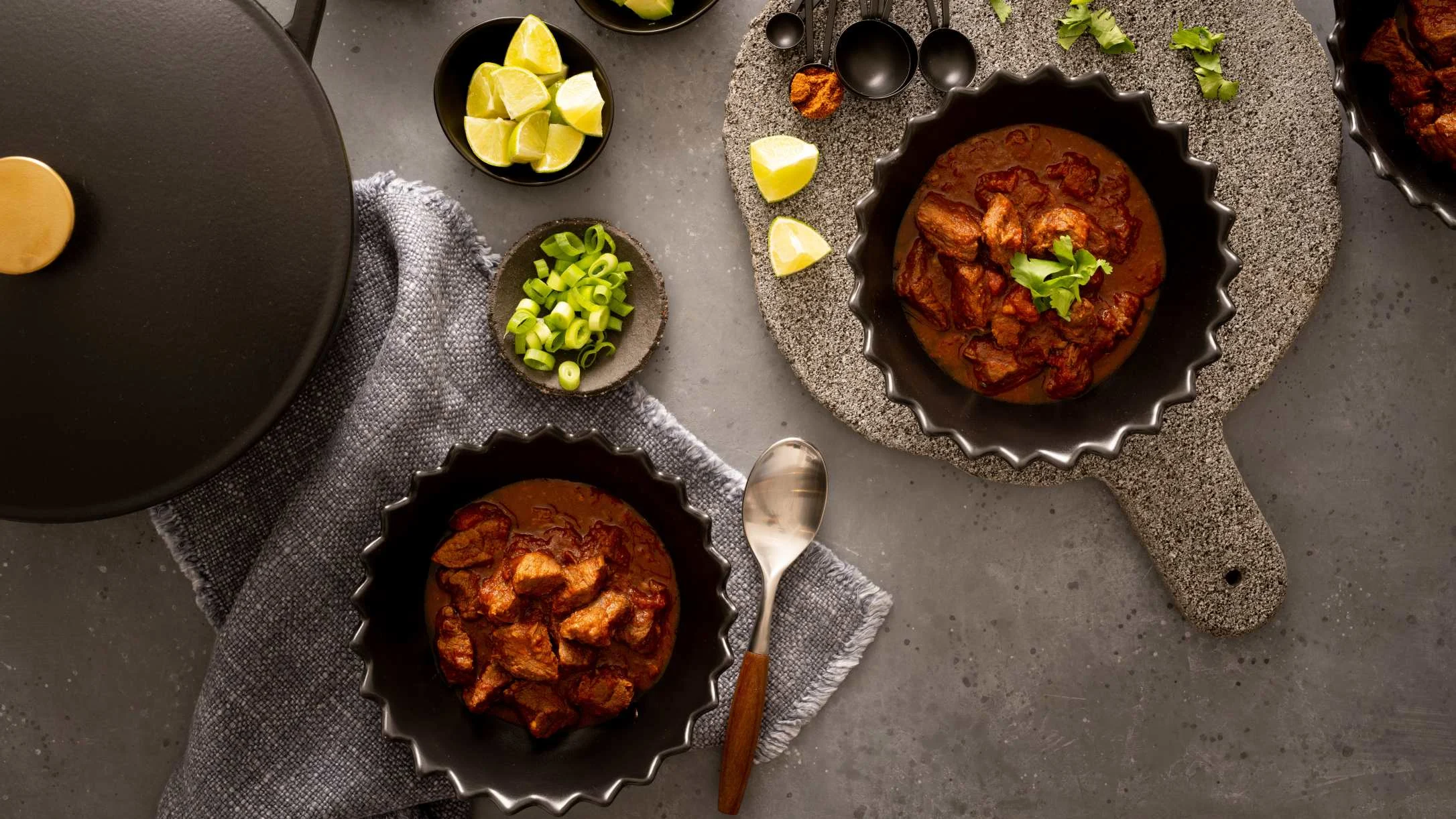 Two dark bowls of meat chili with cilantro, served alongside lime wedges, scallions, and measuring spoons on a gray surface.