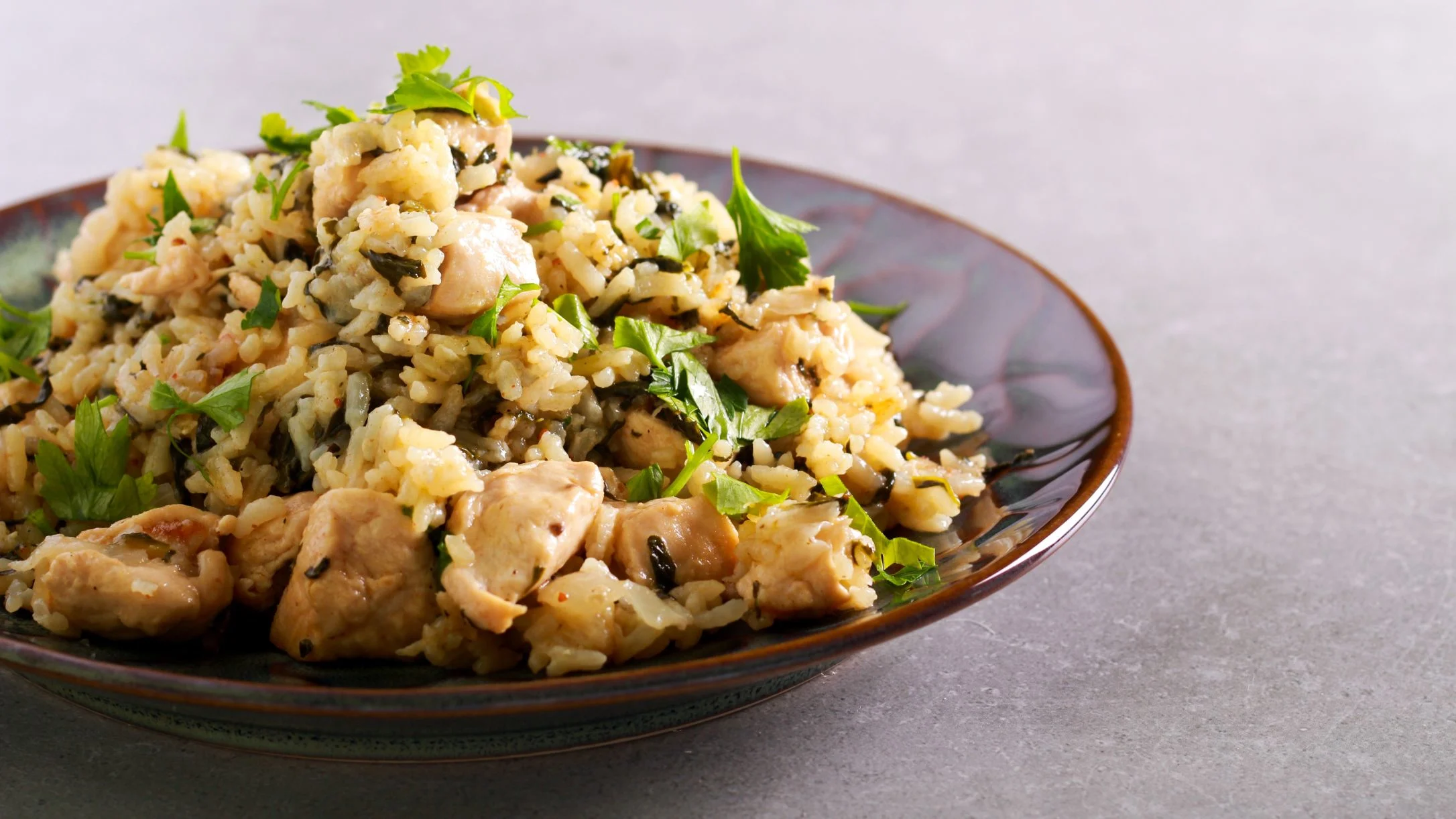A dark ceramic plate piled with chicken and rice garnished with fresh green parsley, set on a gray surface.